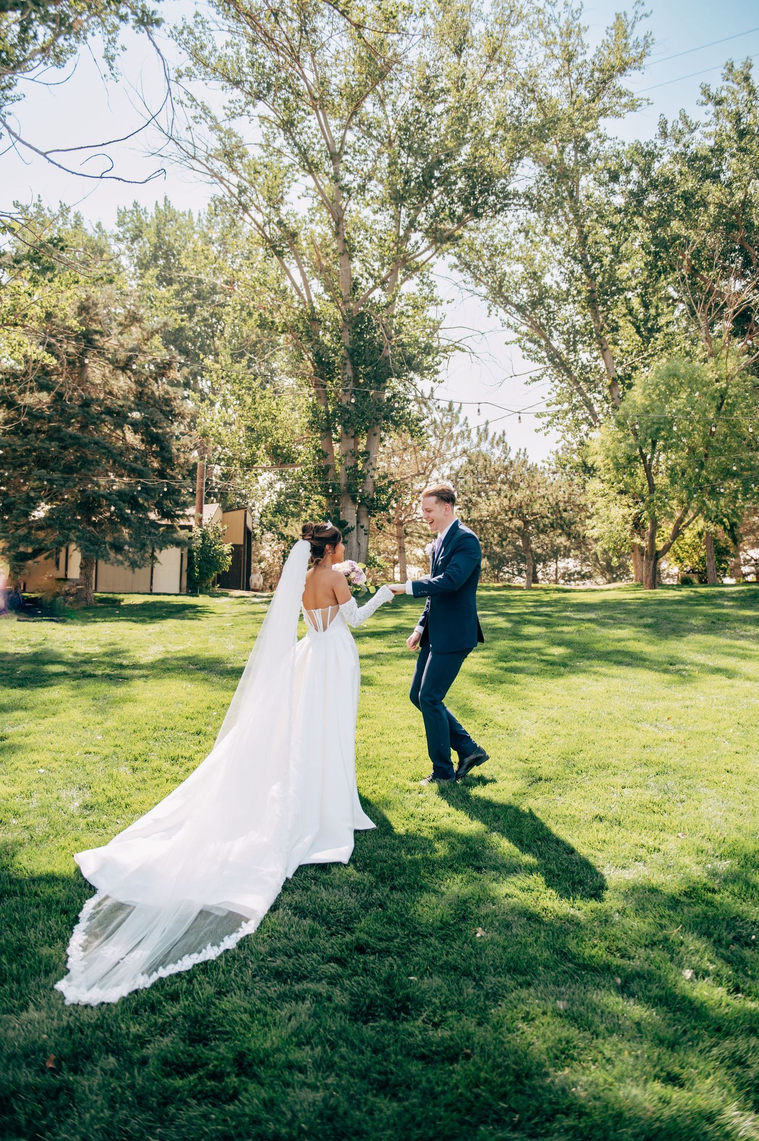 Bride in white gown with long veil and groom in navy suit share a romantic moment on green lawn surrounded by trees.