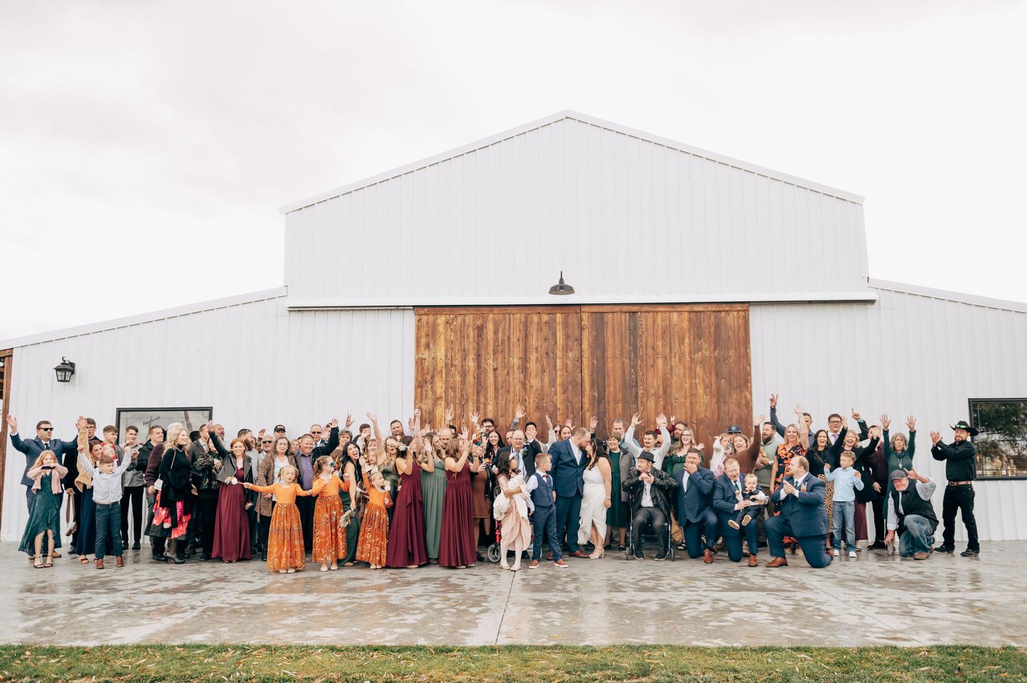 Large group wedding photo outside a modern white barn building with wooden accent wall.