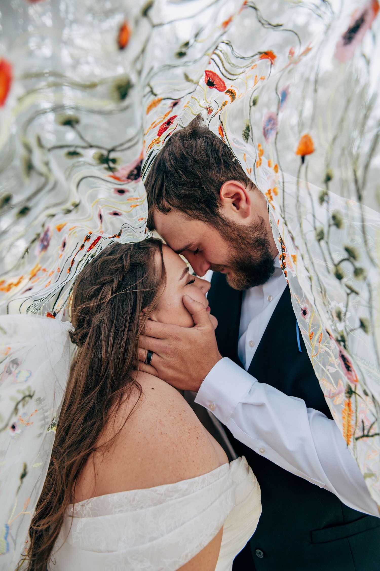 A romantic couple in wedding attire share an intimate moment under a floral veil, groom tenderly holding bride's face.