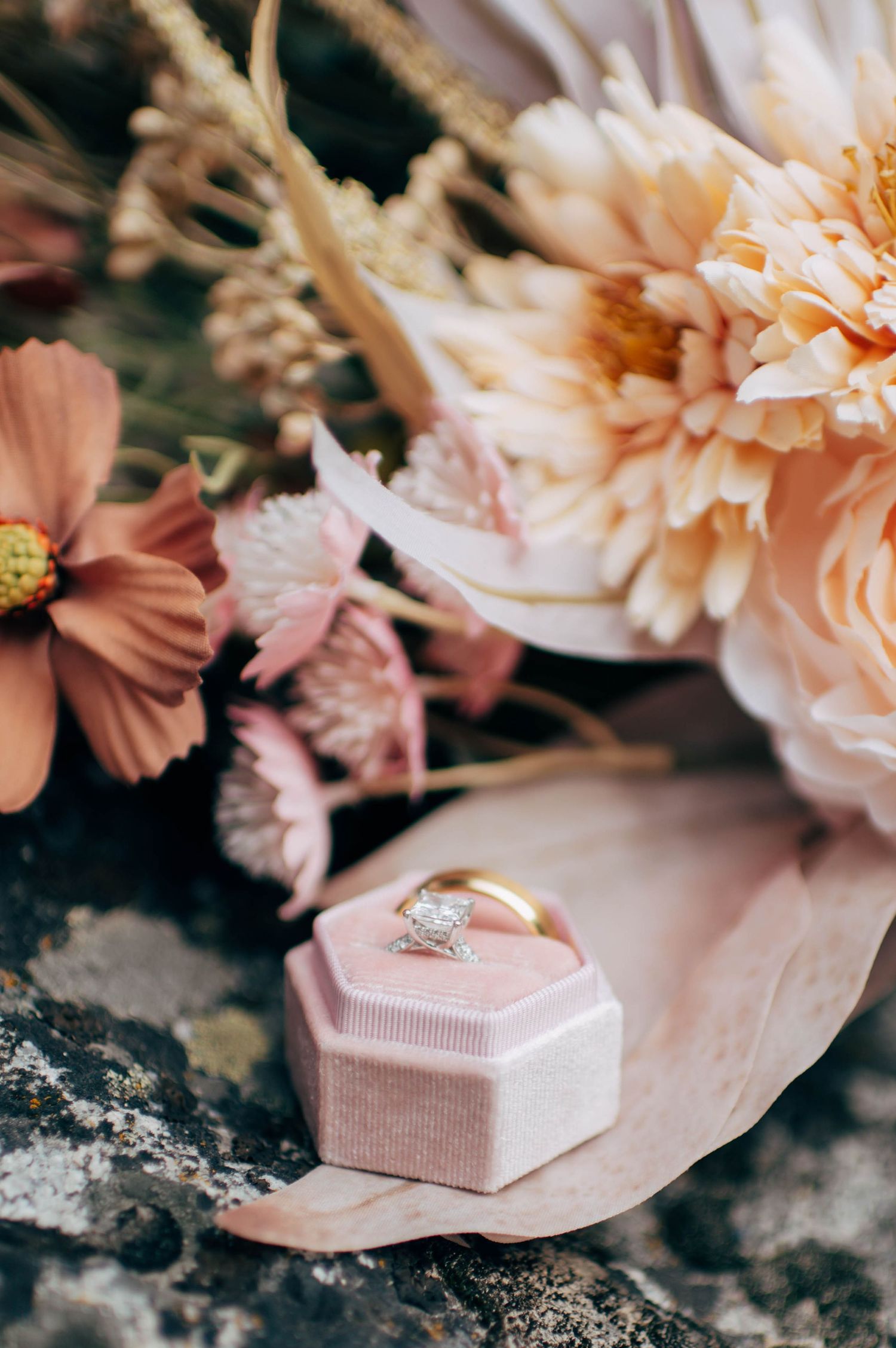 Gold diamond engagement ring in pink velvet box surrounded by blush dahlias and cosmos flowers.
