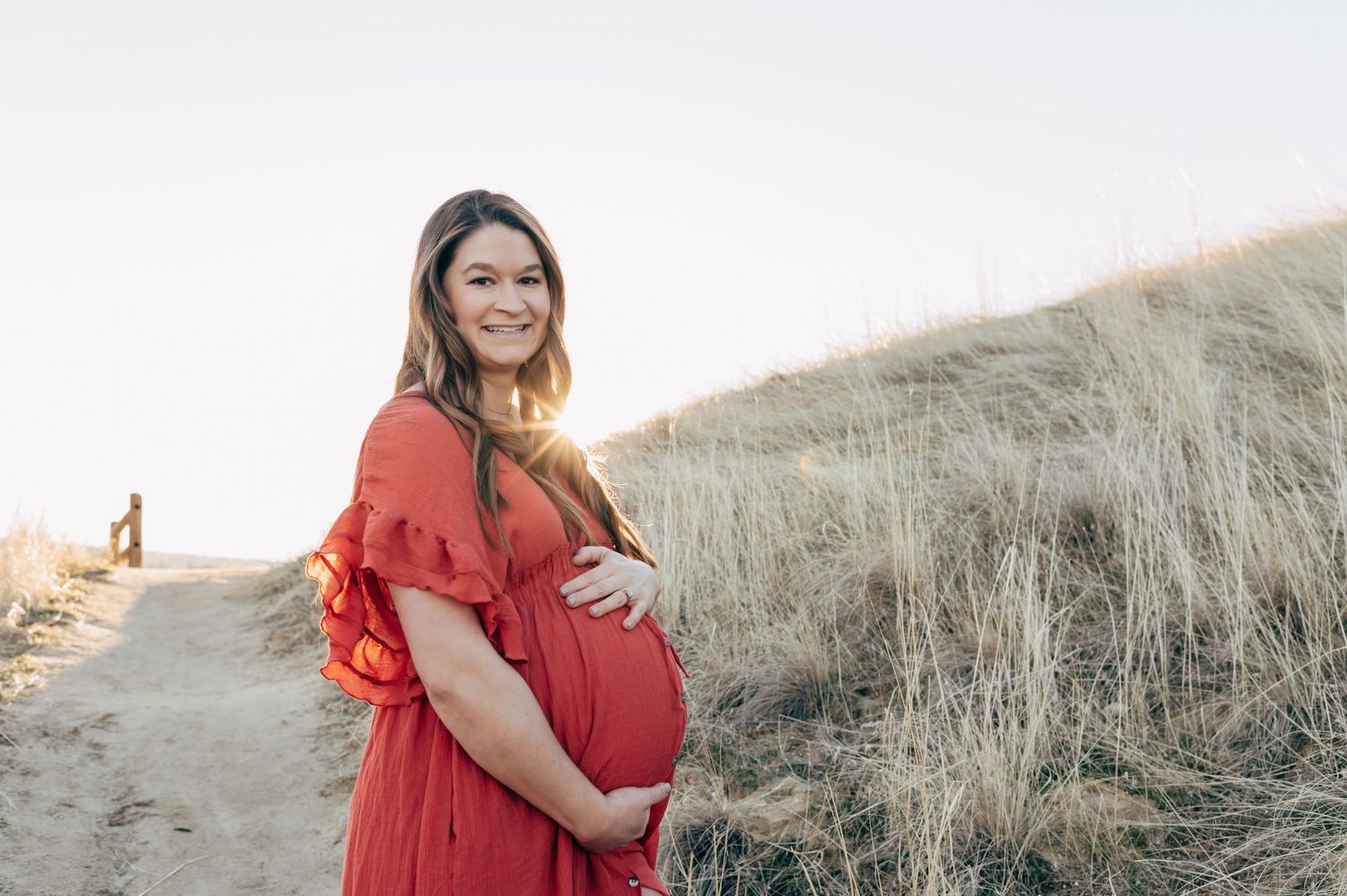 Smiling pregnant woman in red dress cradling baby bump on sandy beach path with golden grass at sunset.