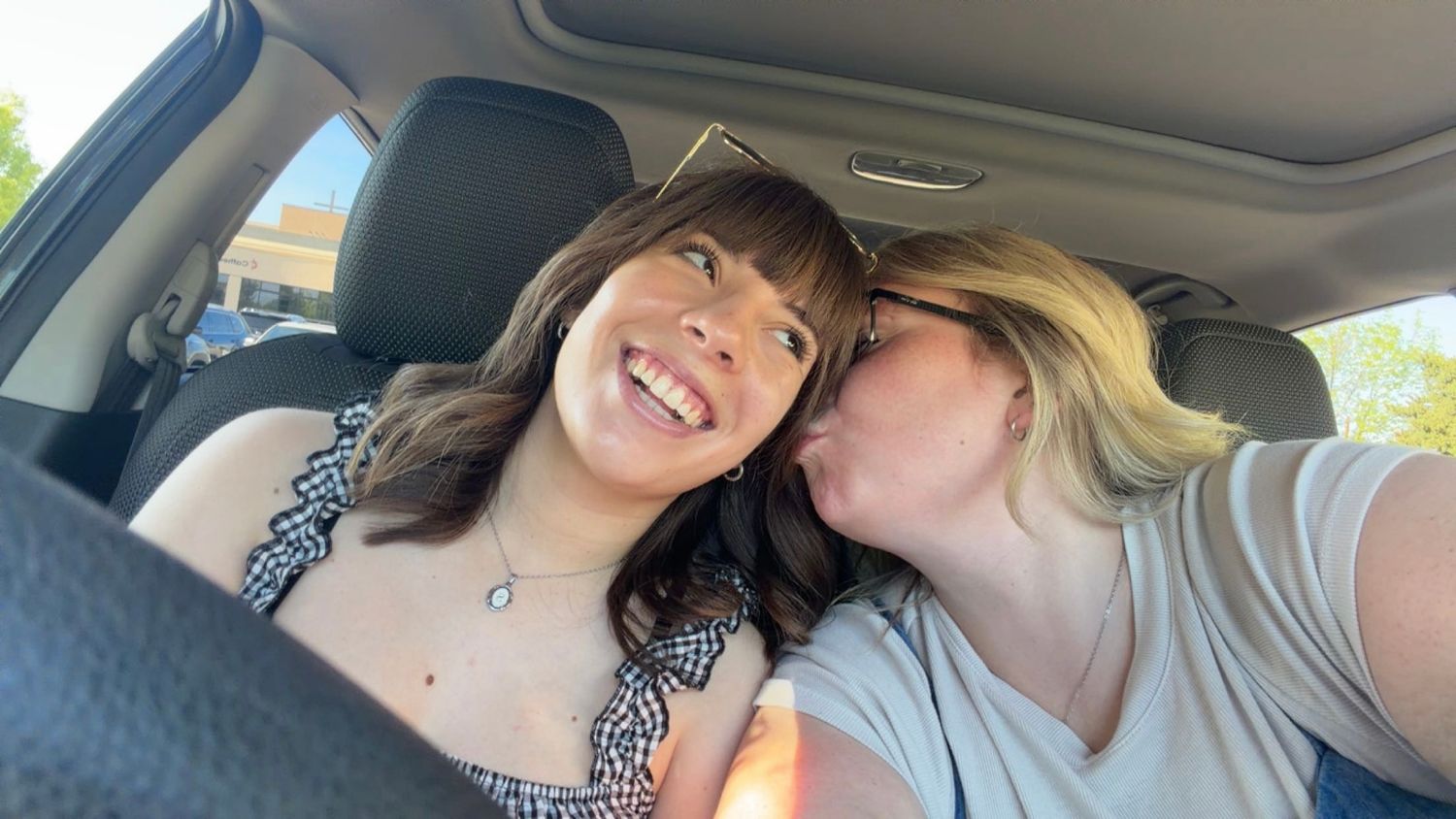 Two women smiling and laughing together inside a car, one giving the other a kiss on the cheek.