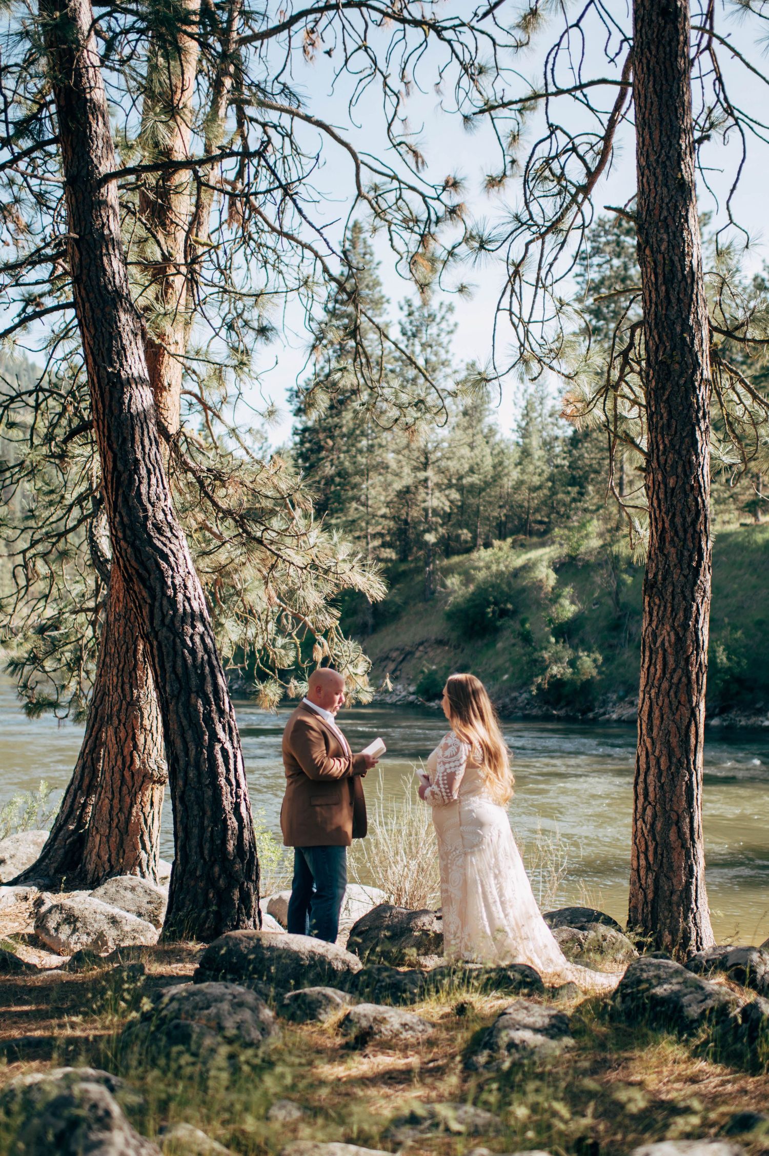 Couple exchanging vows outdoors between pine trees by a river during an intimate elopement ceremony.