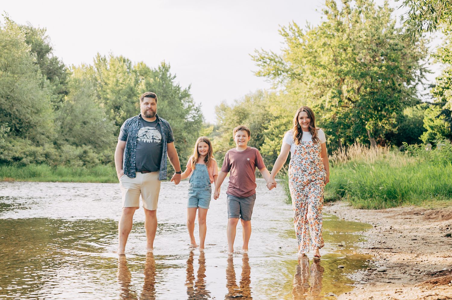 Family of four walking hand-in-hand along a sunny shallow river with lush green trees in the background.