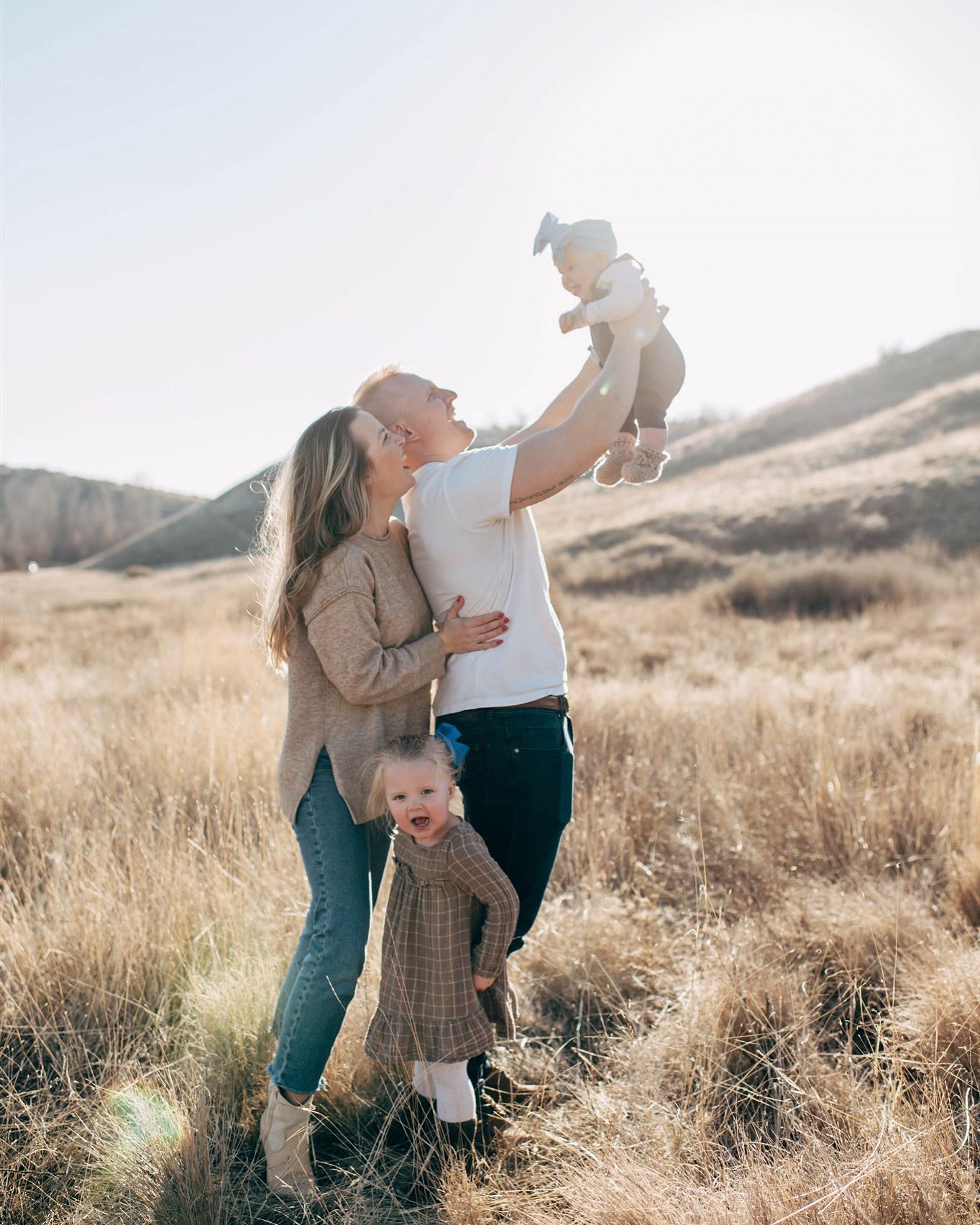 A happy family poses in golden fields, parents kissing while lifting baby and toddler stands nearby.