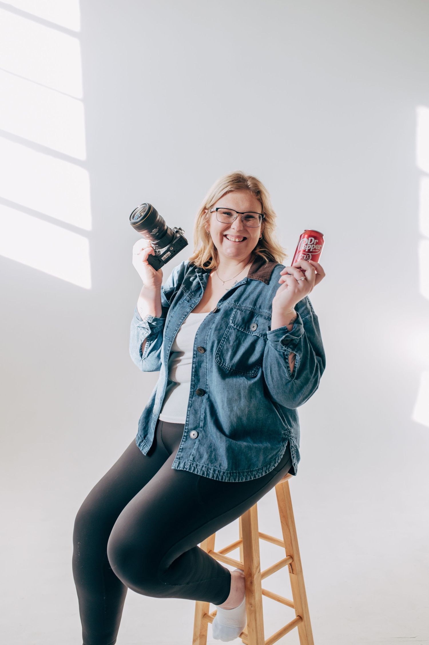 Photographer in denim jacket and black leggings poses with cameras on wooden stool in bright white studio.