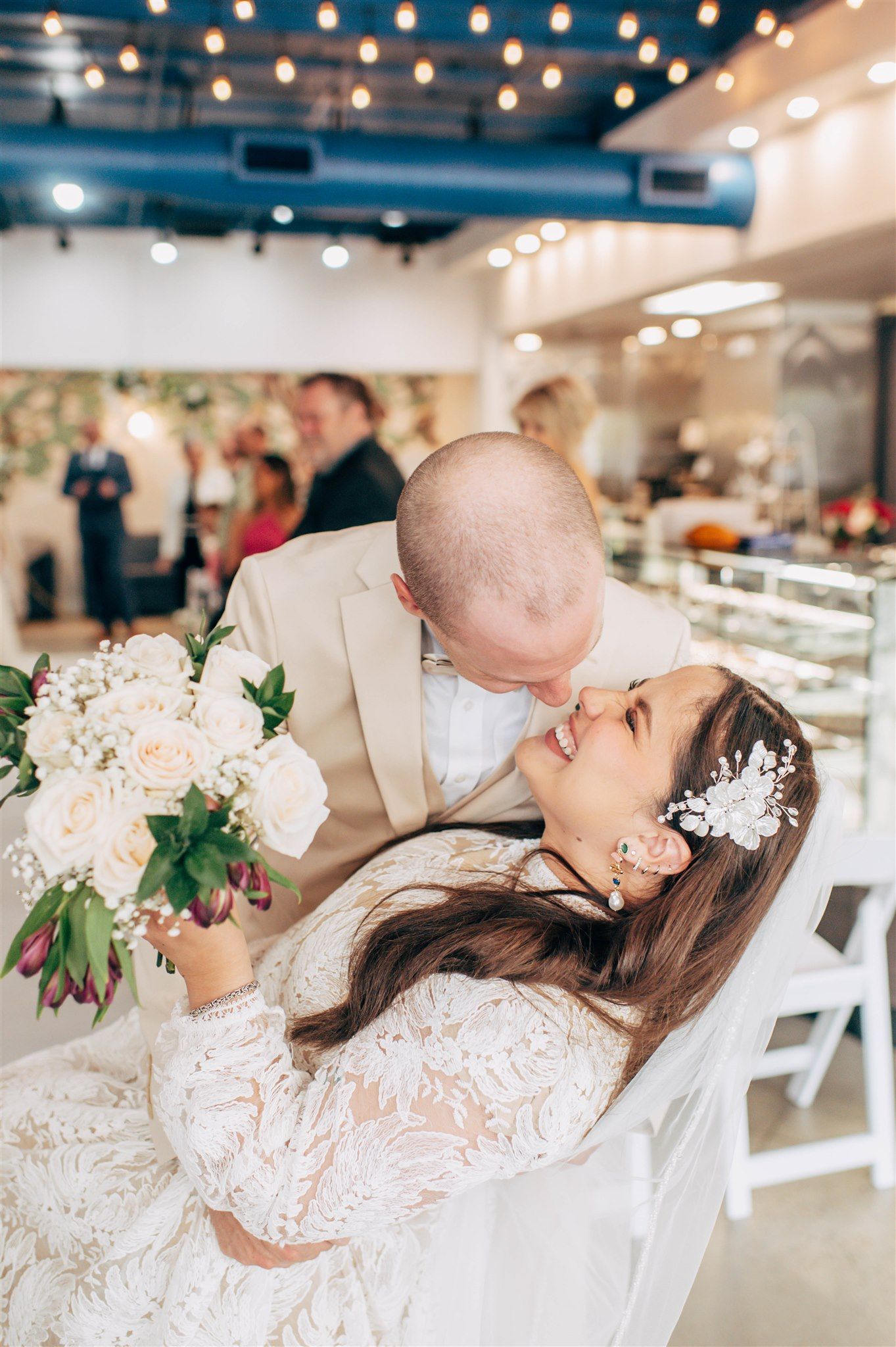 Bride in lace gown holds white roses bouquet as groom in tan suit leans in for a kiss at indoor wedding venue.