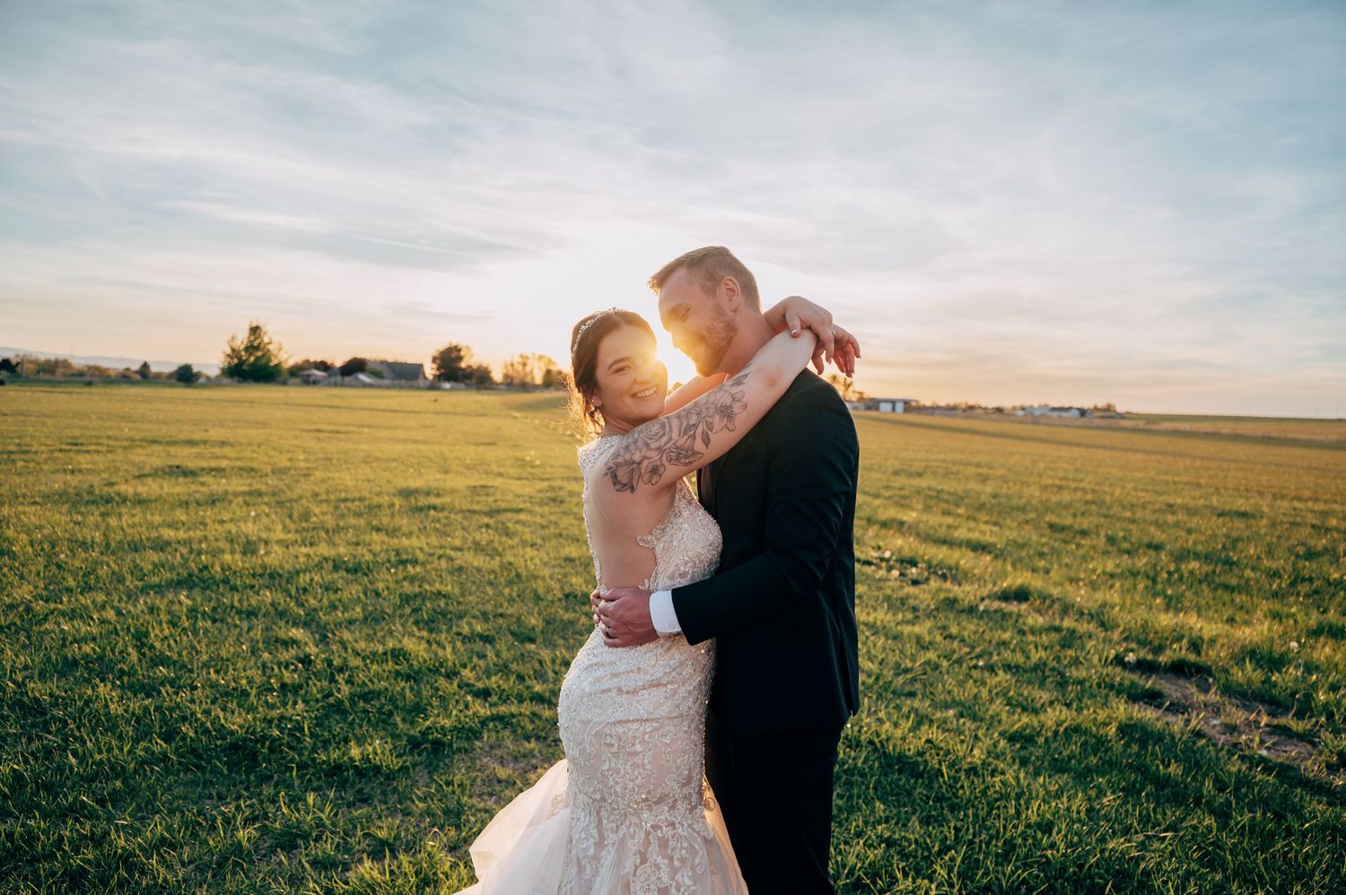 Bride and groom embracing at sunset in a green field, bride in lace gown, groom in dark suit.