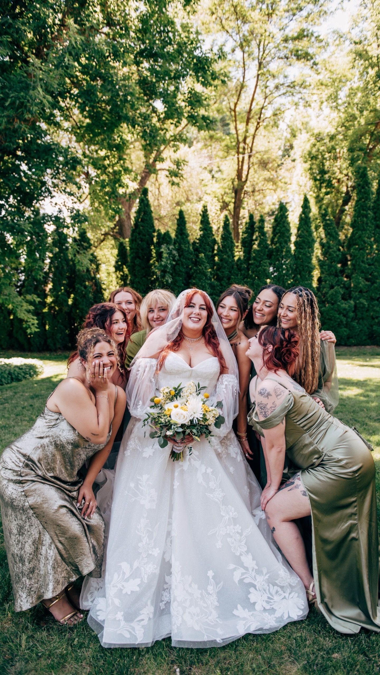 Bridal party in sage dresses gathered around brides in white gowns on sunny lawn.
