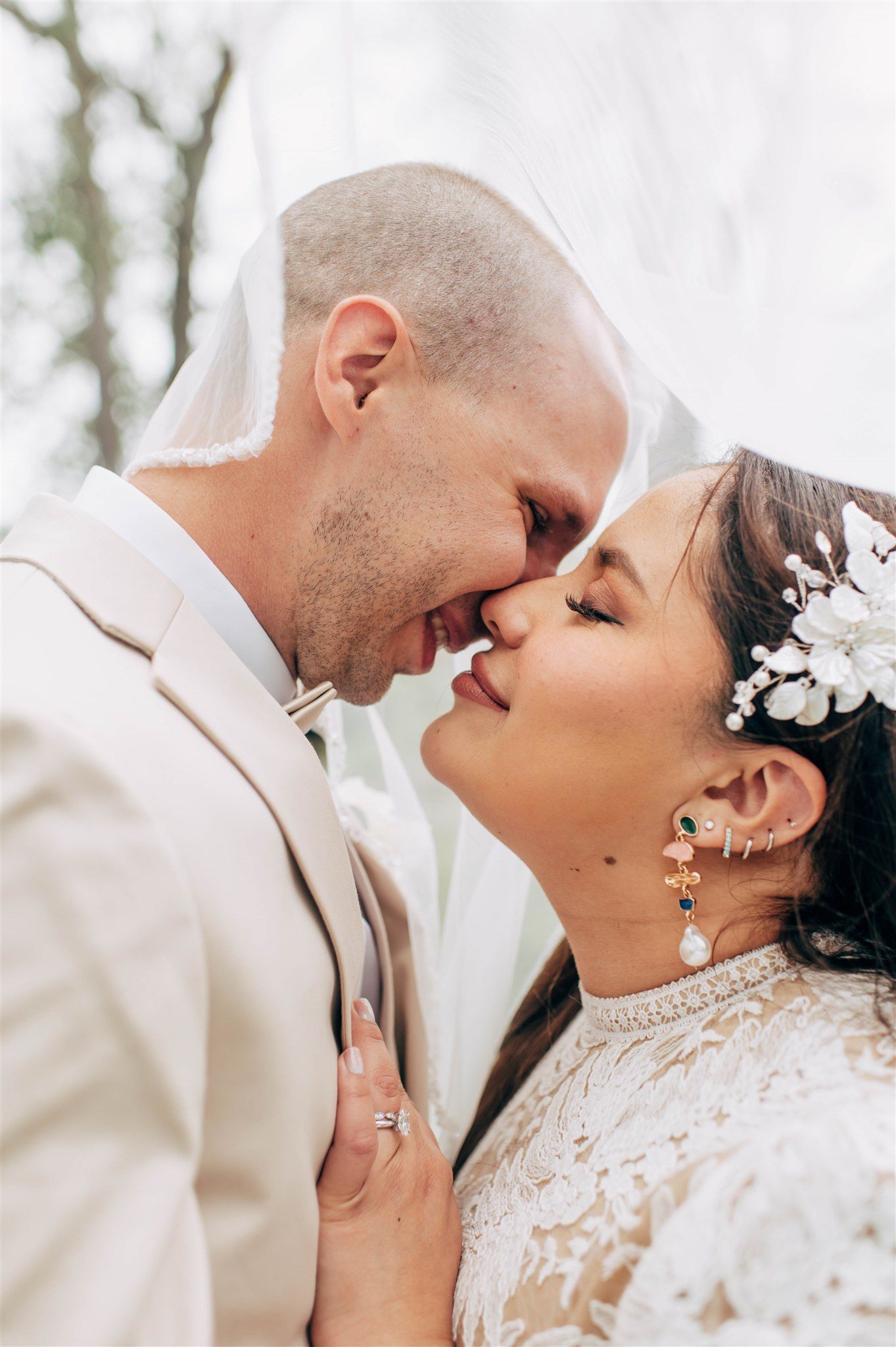 A bride and groom share a romantic almost-kiss under a veil, bride wearing floral hair accessories and colorful earrings.