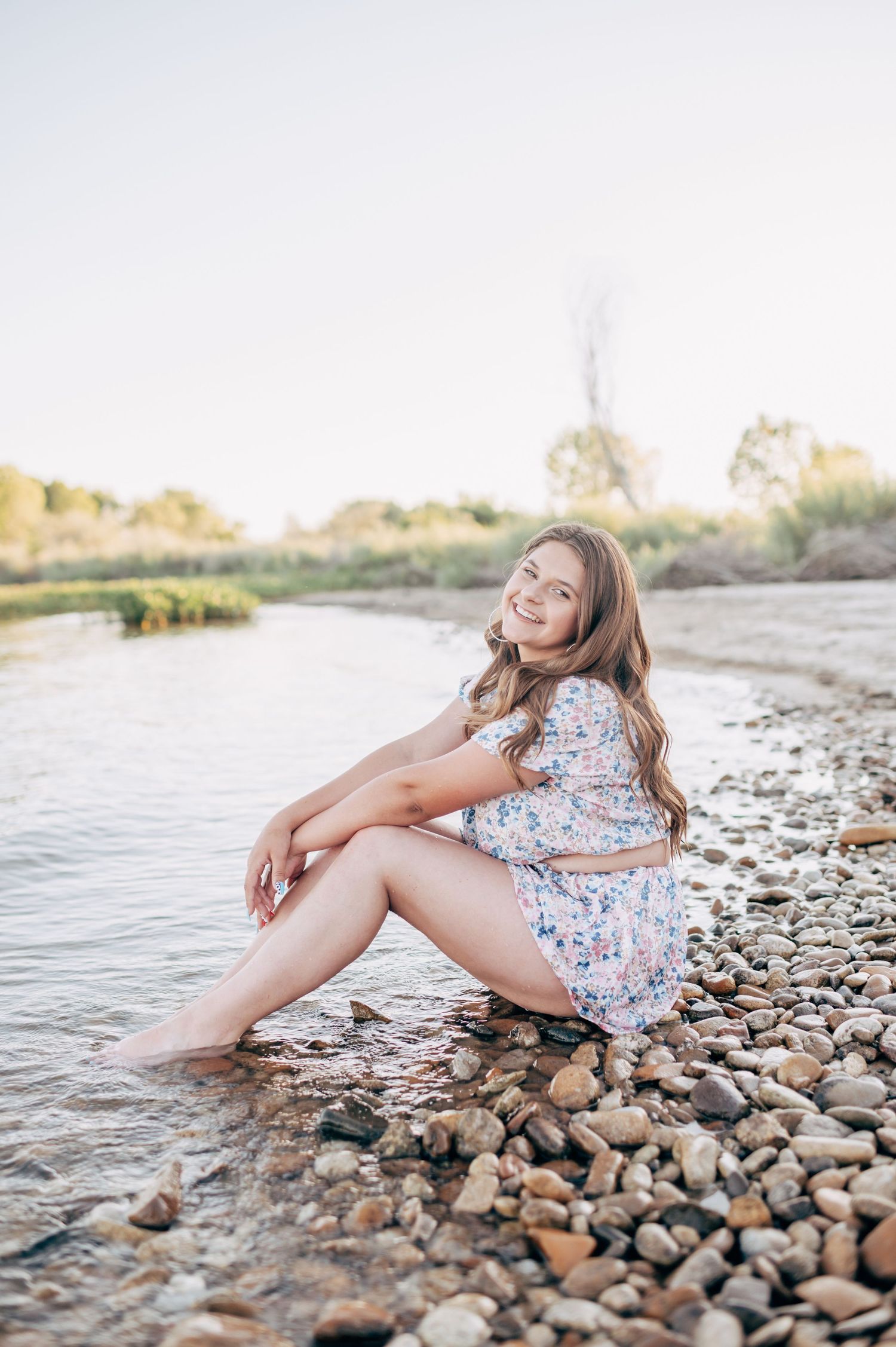Young woman in floral dress sitting barefoot on rocky riverbank, smiling outdoors in warm natural light.
