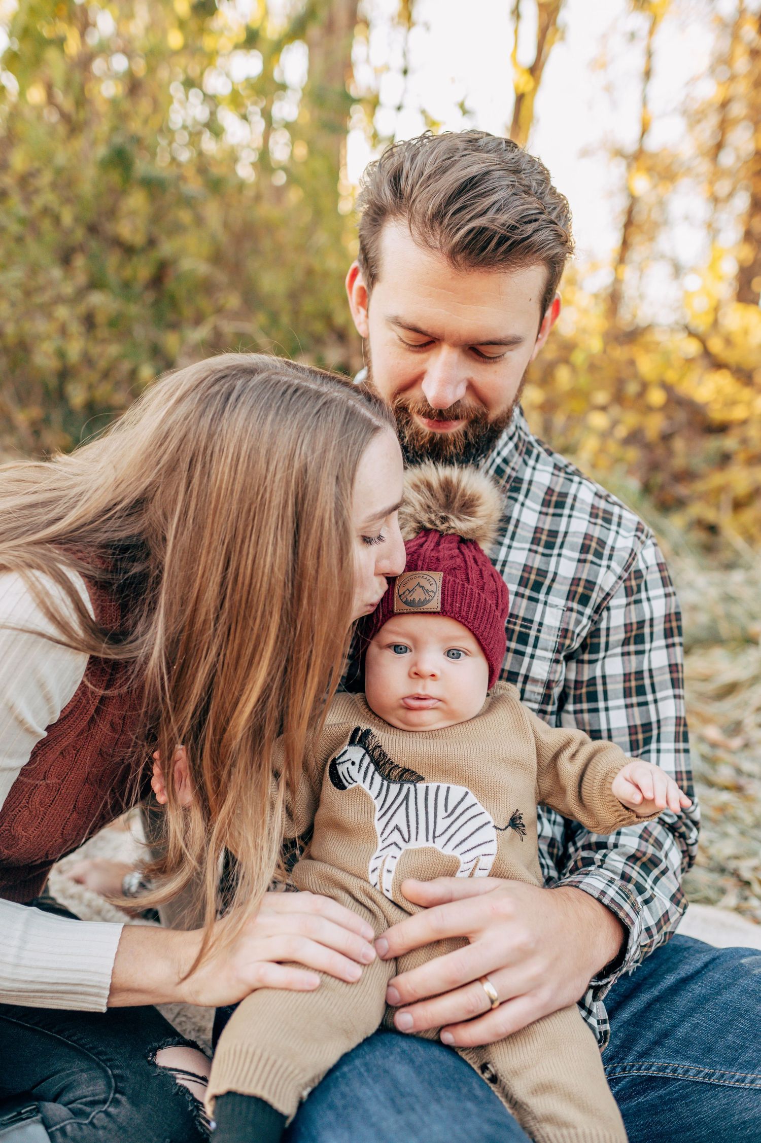Young couple kissing newborn baby wearing red knit hat outdoors in autumn forest setting.