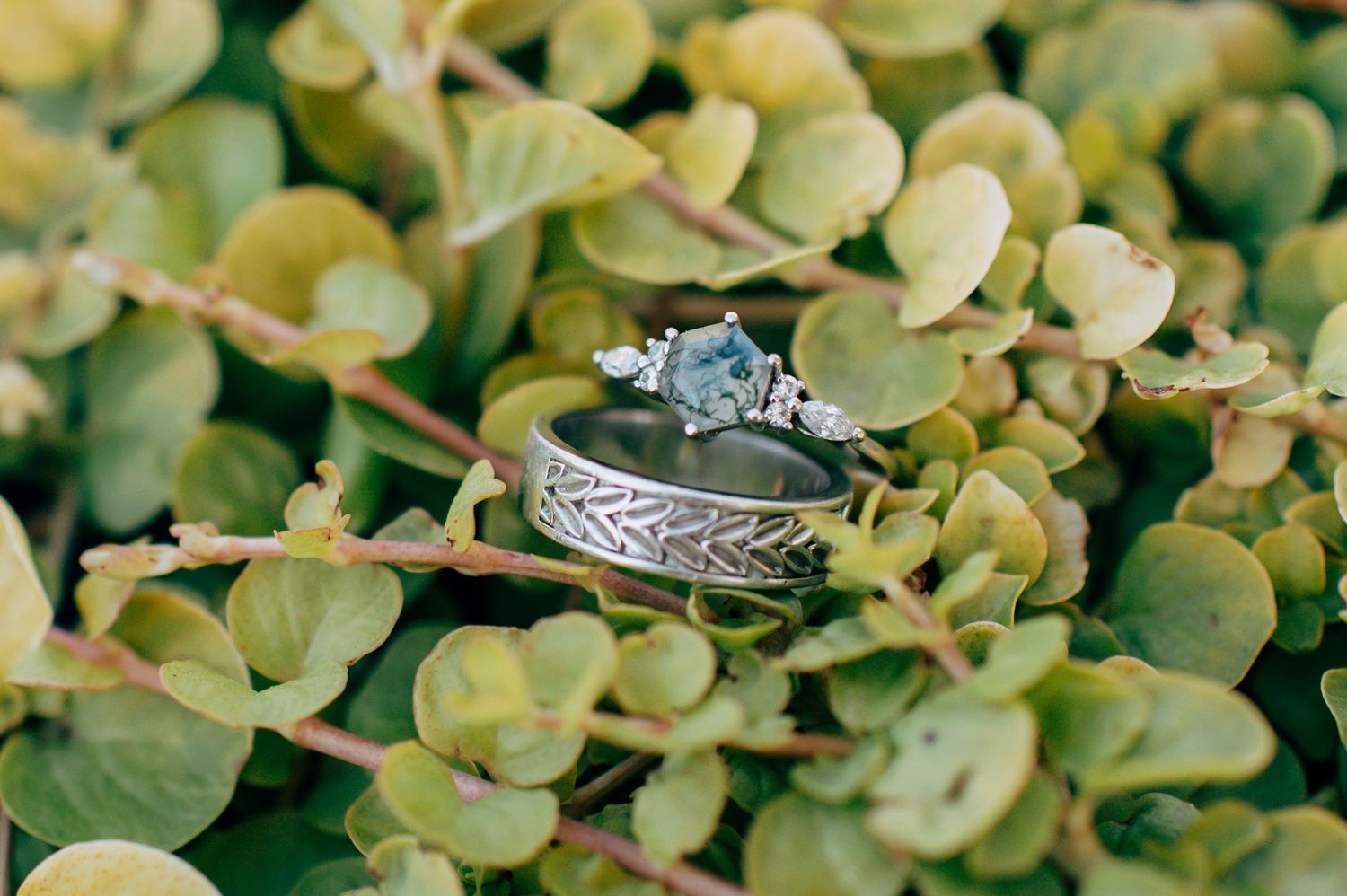 Two elegant wedding rings resting on green foliage, featuring an engraved band and a blue gemstone engagement ring.