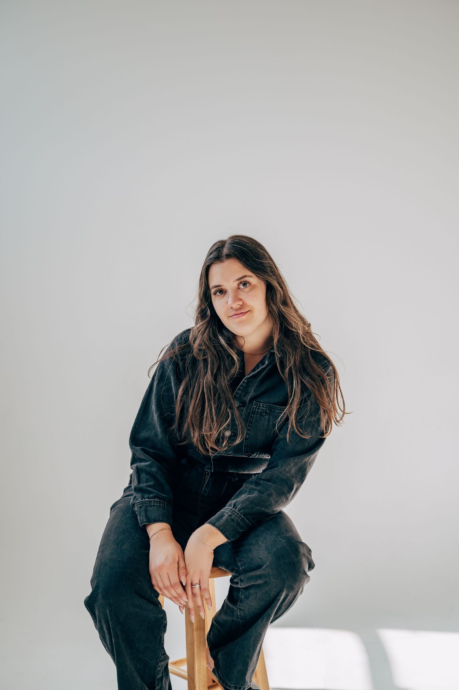A young woman with long brown hair sits on a wooden stool wearing a black denim outfit against a white background.