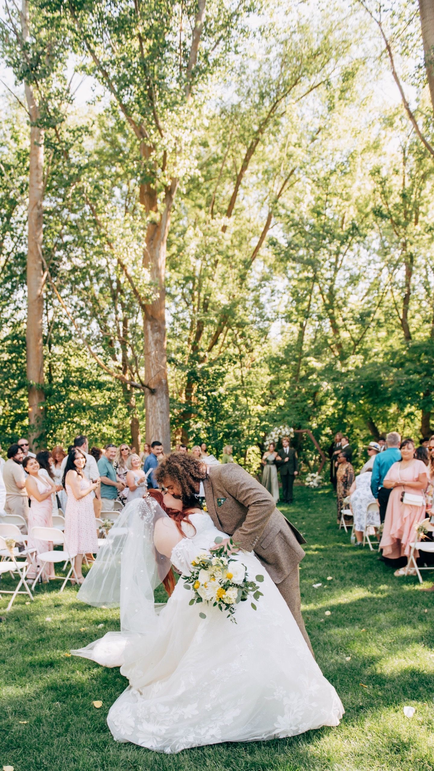 Wedding ceremony dip kiss with guests clapping in sunlit outdoor garden setting.