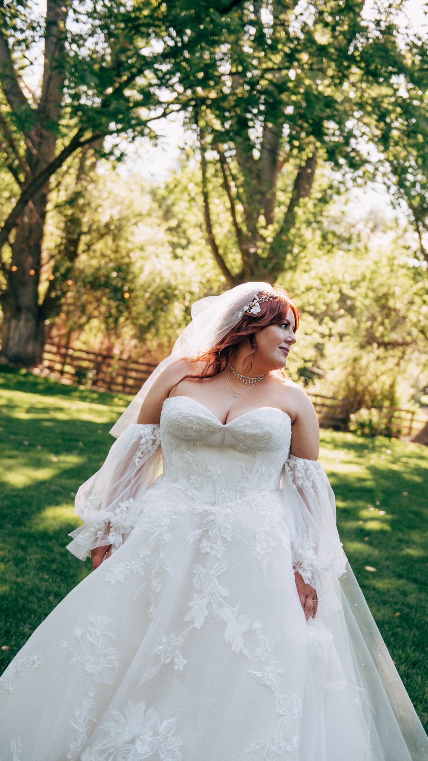Bride in an elegant white lace wedding gown poses in a sunlit garden setting.