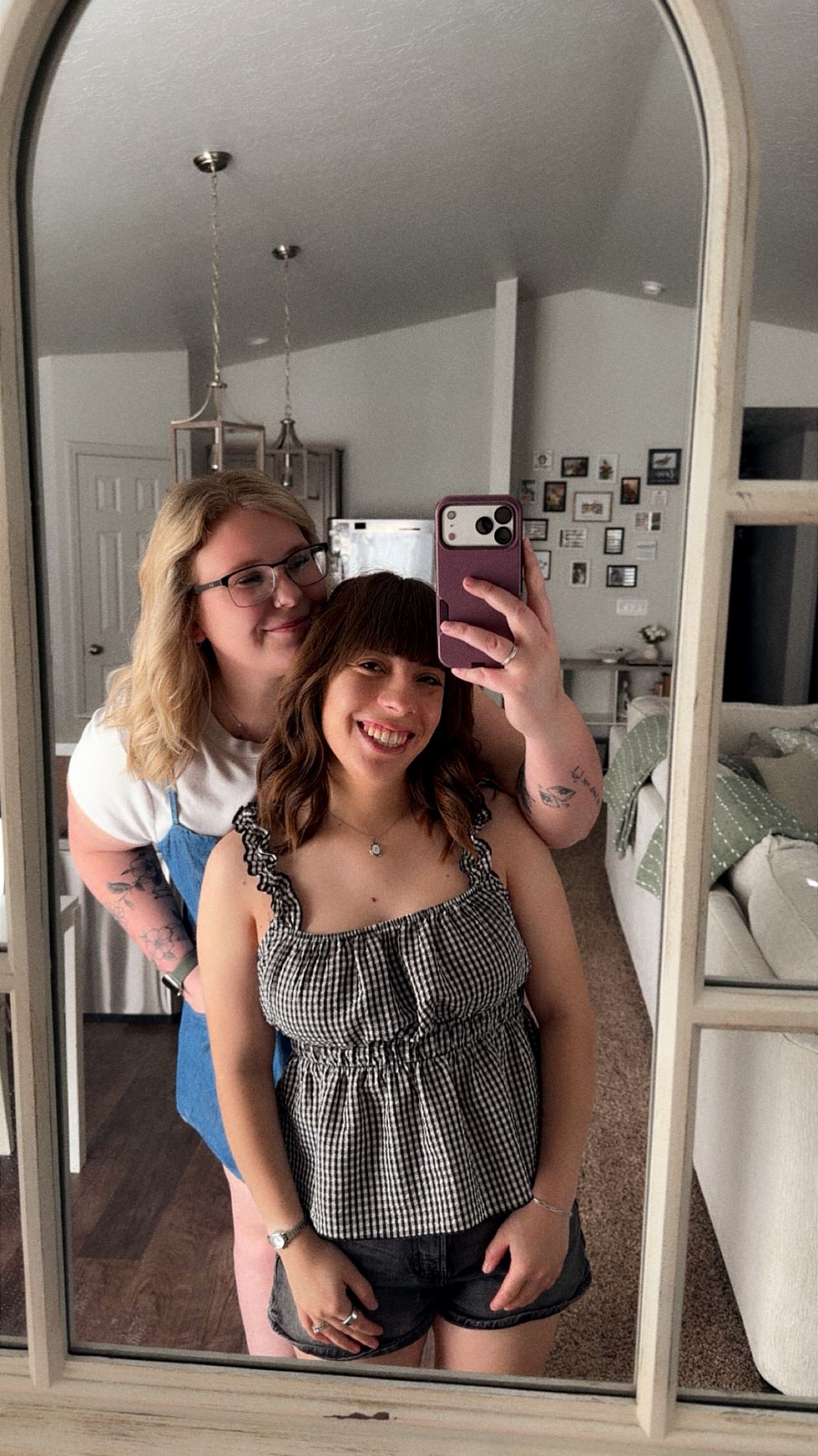 Two young women smiling together in a mirror selfie, one wearing glasses and a white top, the other in a gingham top.