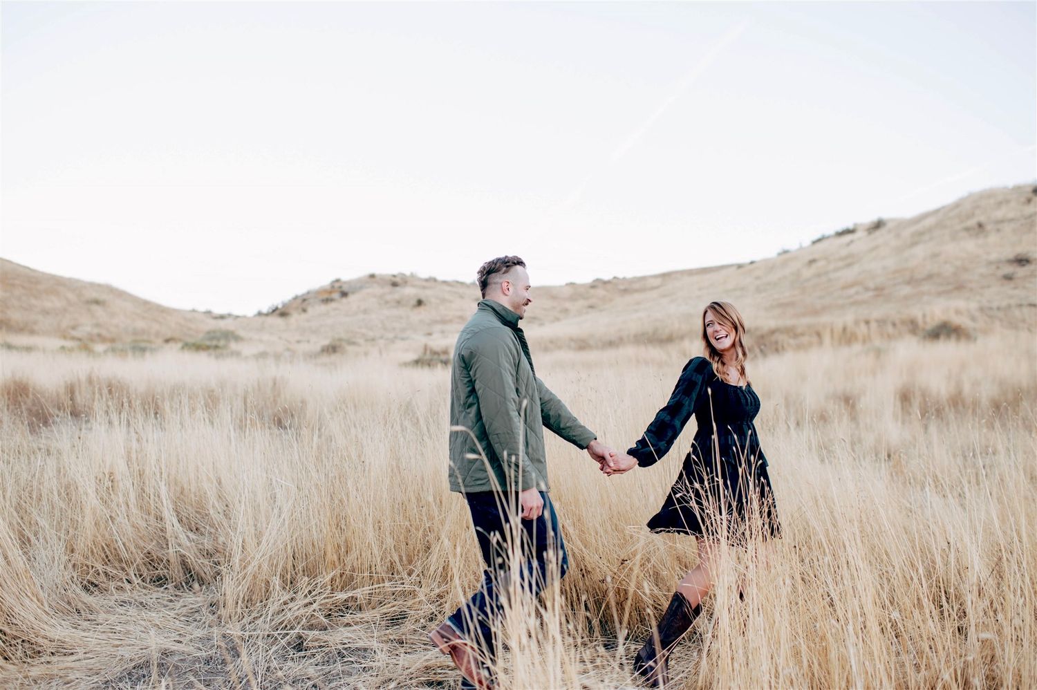 A couple holds hands while walking through tall golden grass in a mountainous landscape.