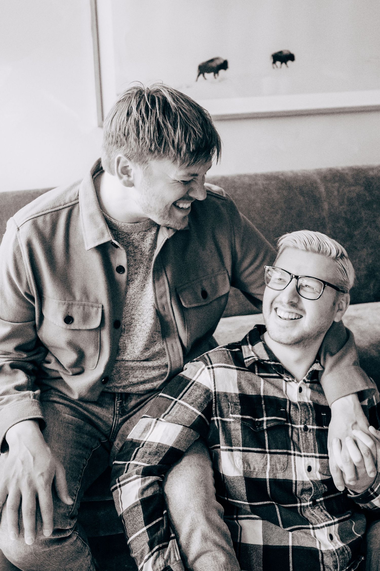 Two young men laughing together on a couch, one wearing glasses and plaid shirt, black and white photo.