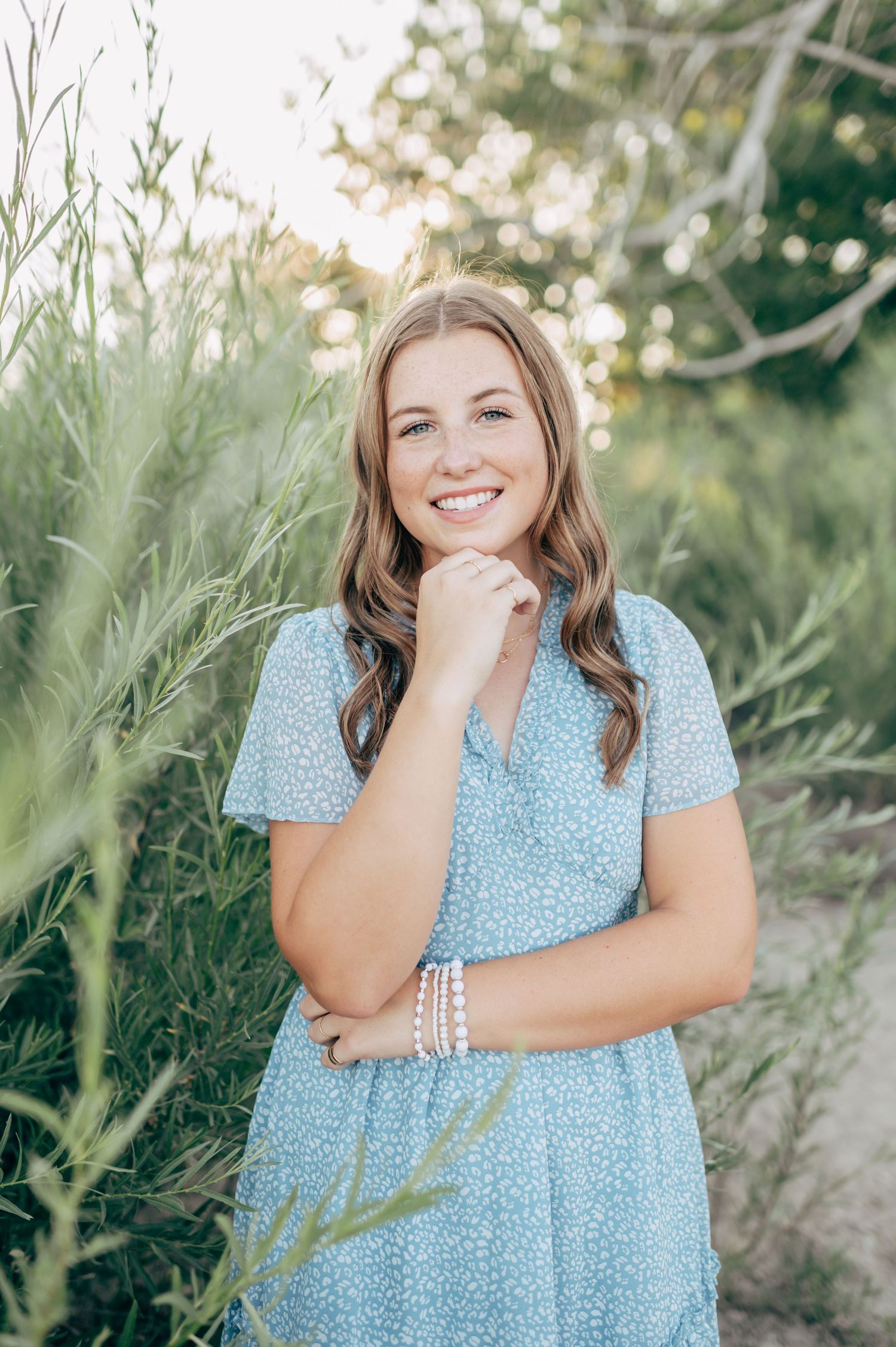 Young woman in light blue floral dress smiling outdoors among green foliage in a bright natural setting.