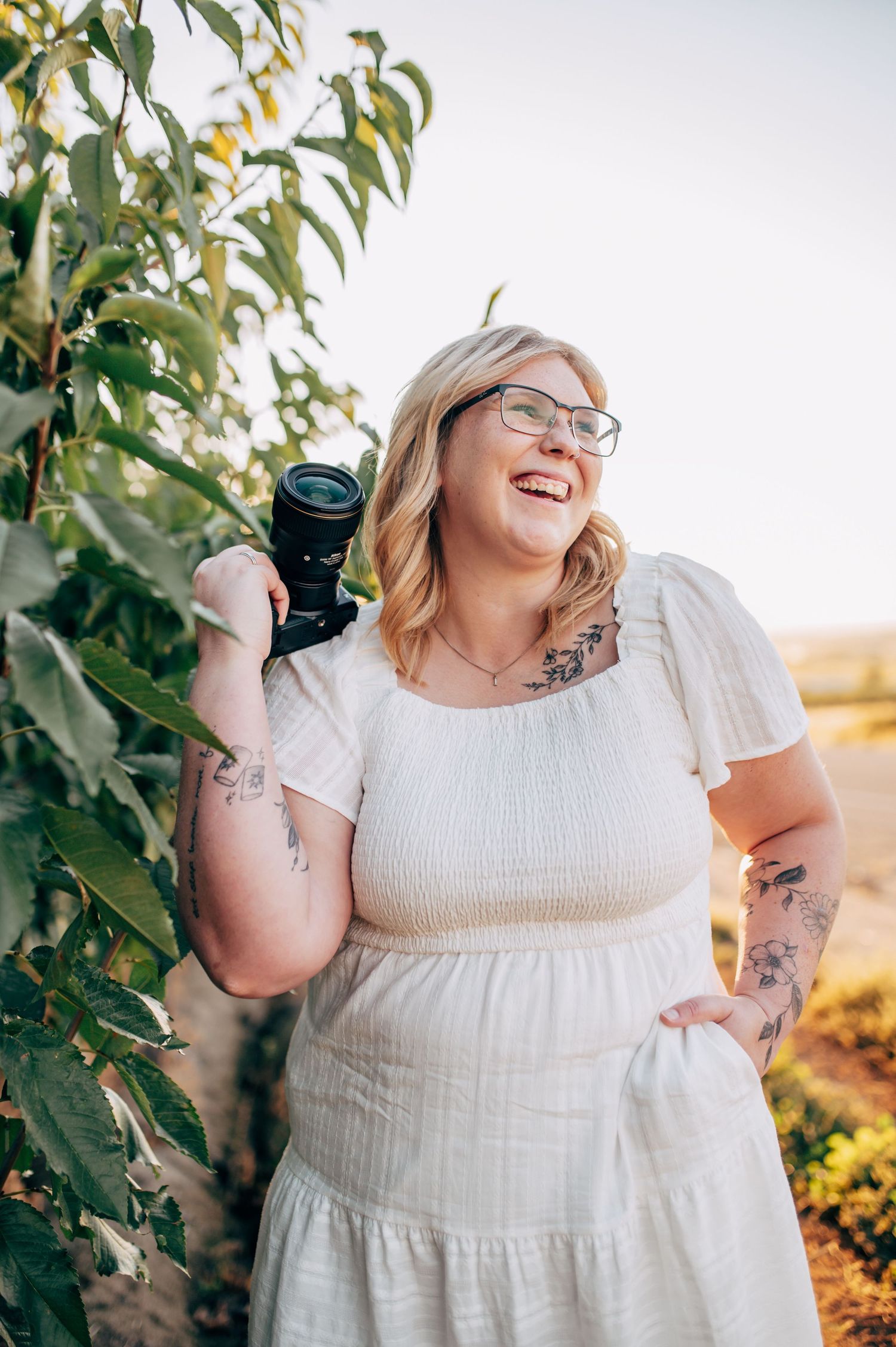 A person in a white dress poses joyfully with a camera during a golden hour photoshoot outdoors.