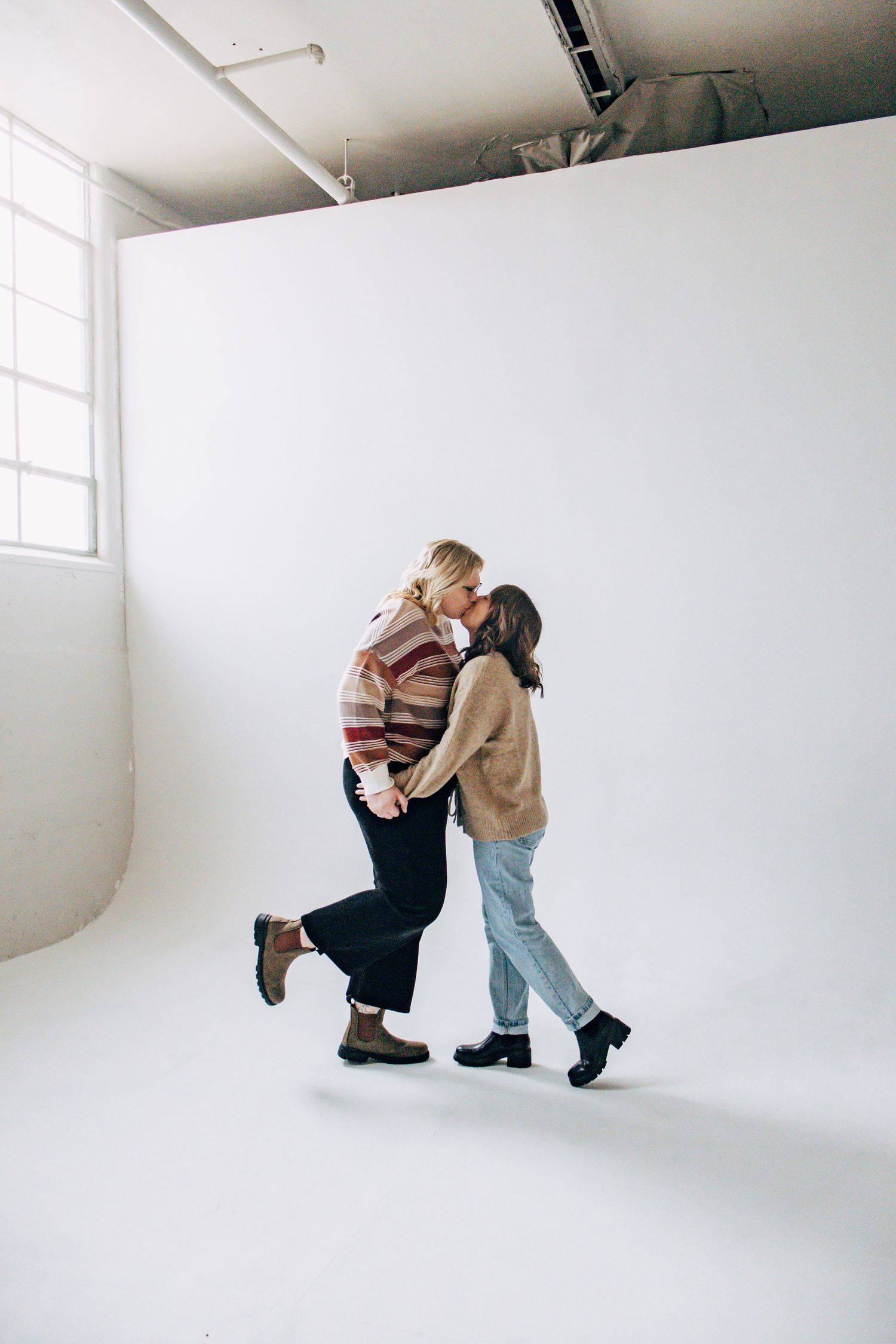 Two people in casual sweaters embrace while dancing together in a minimalist white studio space.