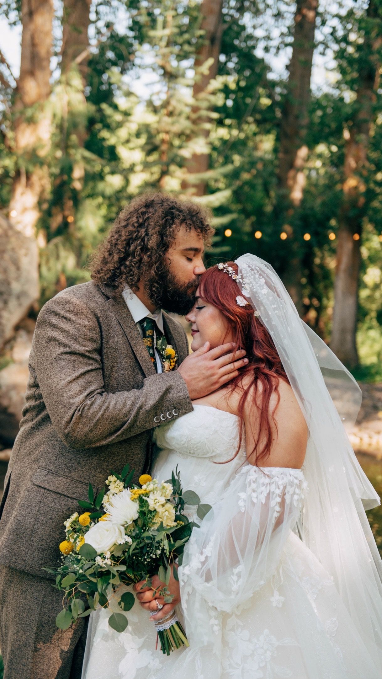 Tender wedding photo of couple embracing under string lights in woodland setting.