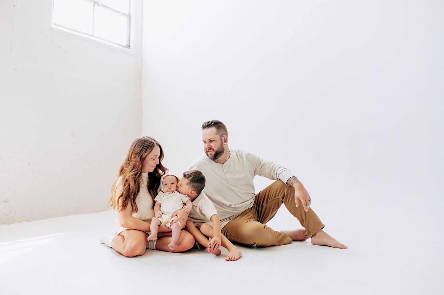 Family of four sitting together on white studio floor, parents smiling at toddler and baby in casual lifestyle portrait.