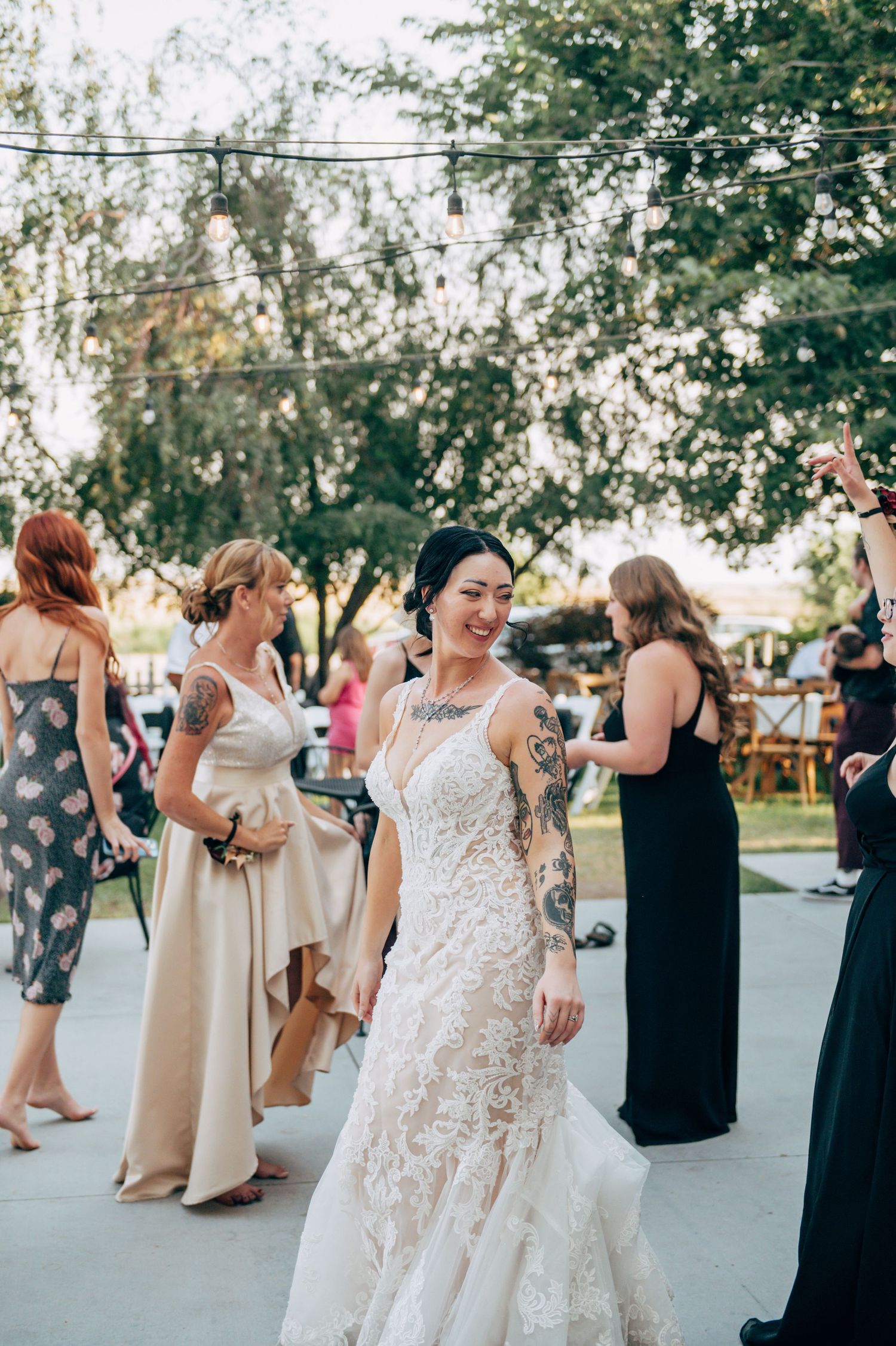Tattooed bride in lace wedding gown smiling at outdoor reception with guests in formal attire.