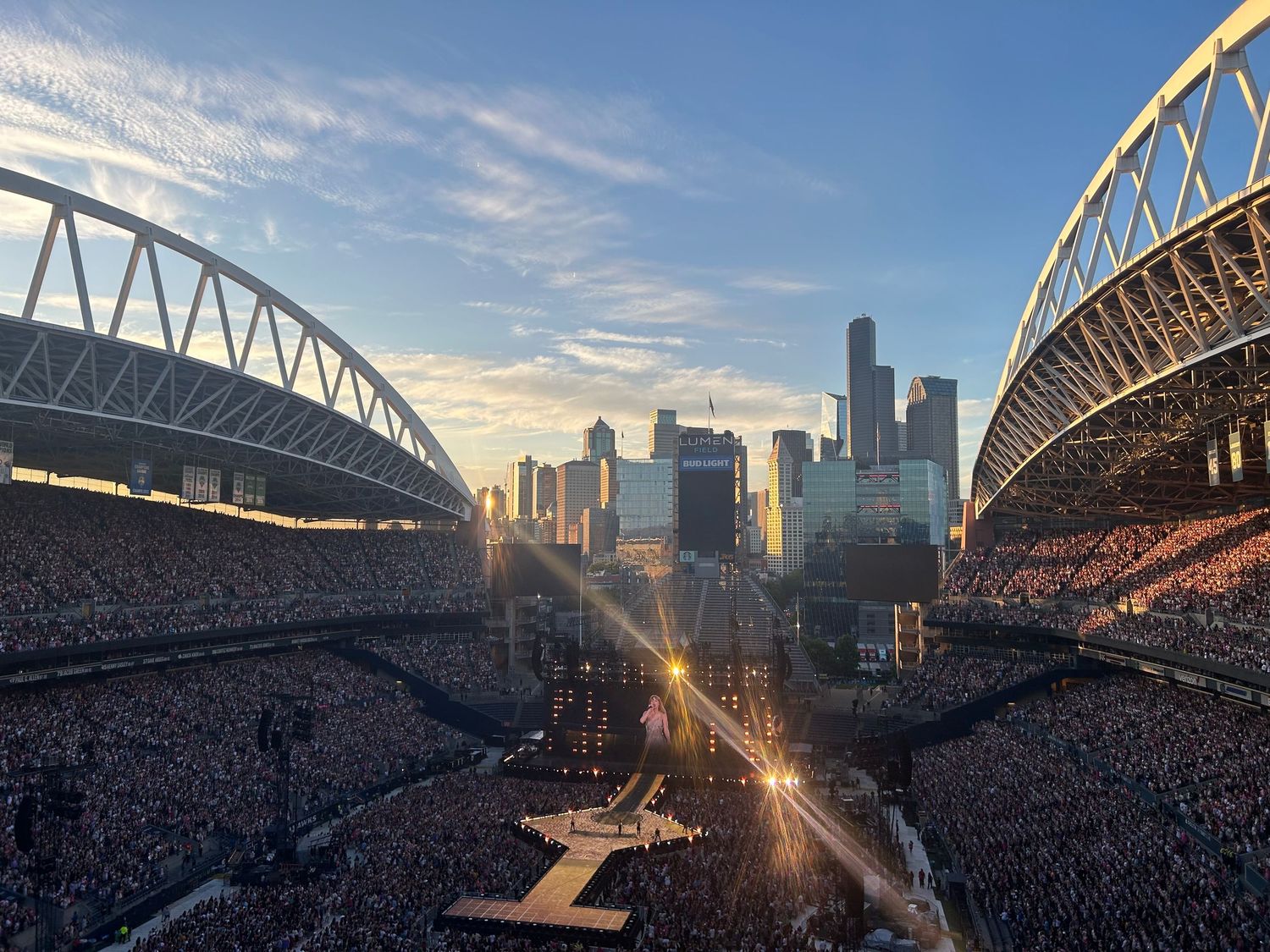 Massive outdoor concert at Seattle stadium with city skyline at sunset, packed crowd and bright stage lights illuminating the venue.