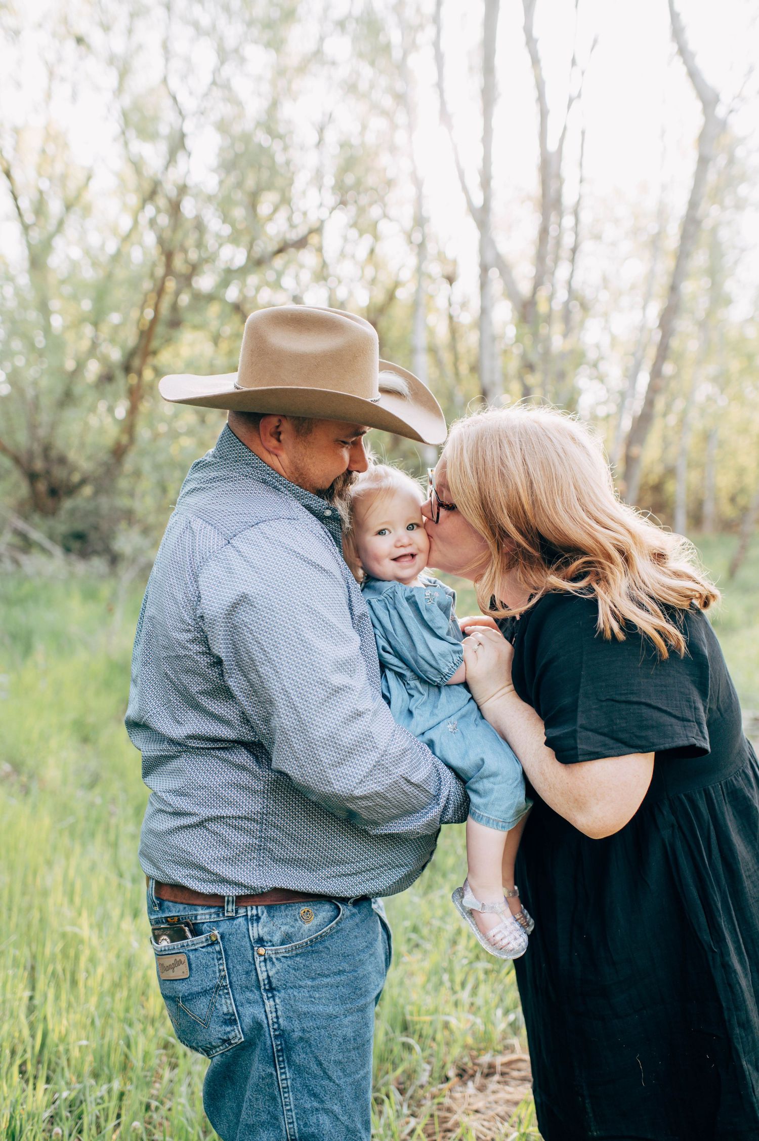 A couple in a green field kisses their smiling toddler, the man wearing a cowboy hat and jeans.