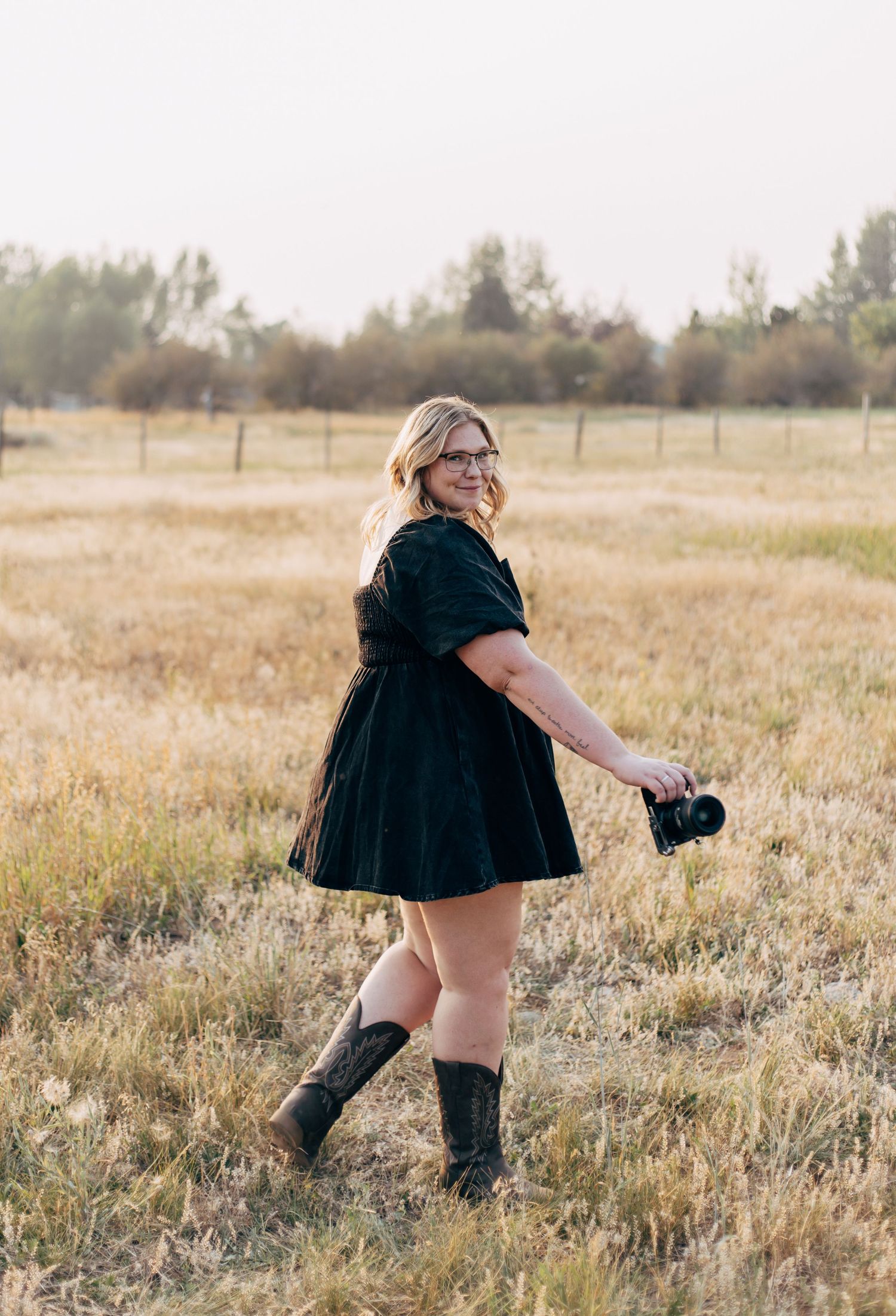 Woman in black dress and cowboy boots holding a camera, standing in a golden field with trees in the background.