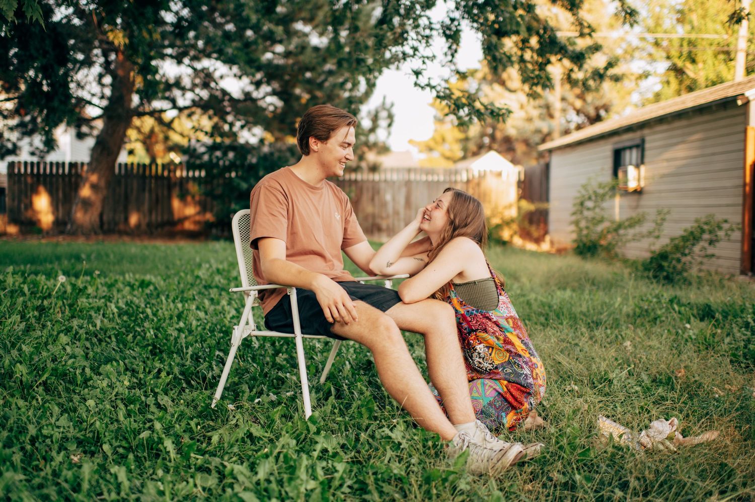 A young couple shares a joyful moment in a sunny backyard, with the girl sitting on the grass beside a boy on a chair.