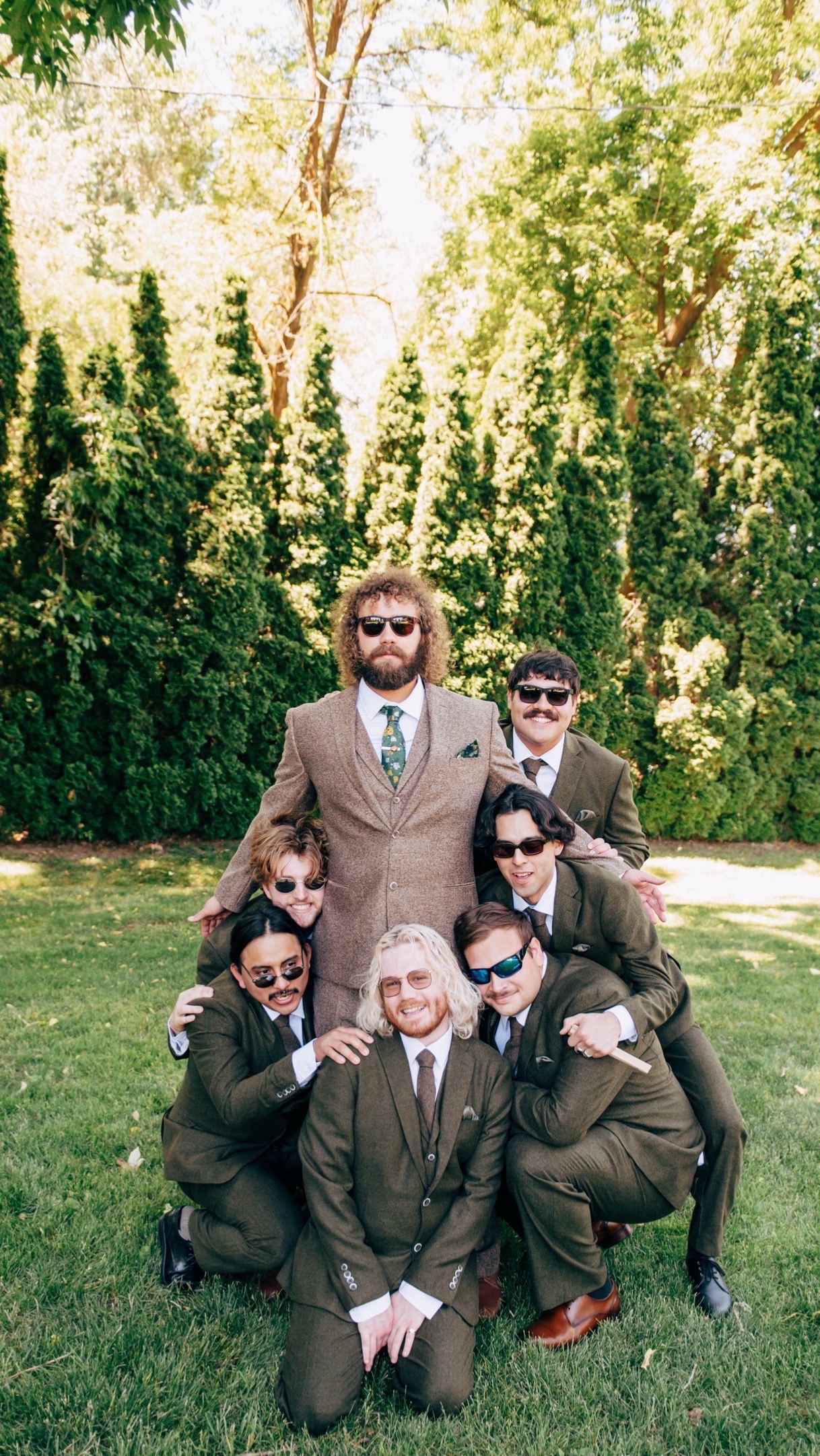 Groomsmen in brown suits and sunglasses posing playfully on grass for wedding photos.
