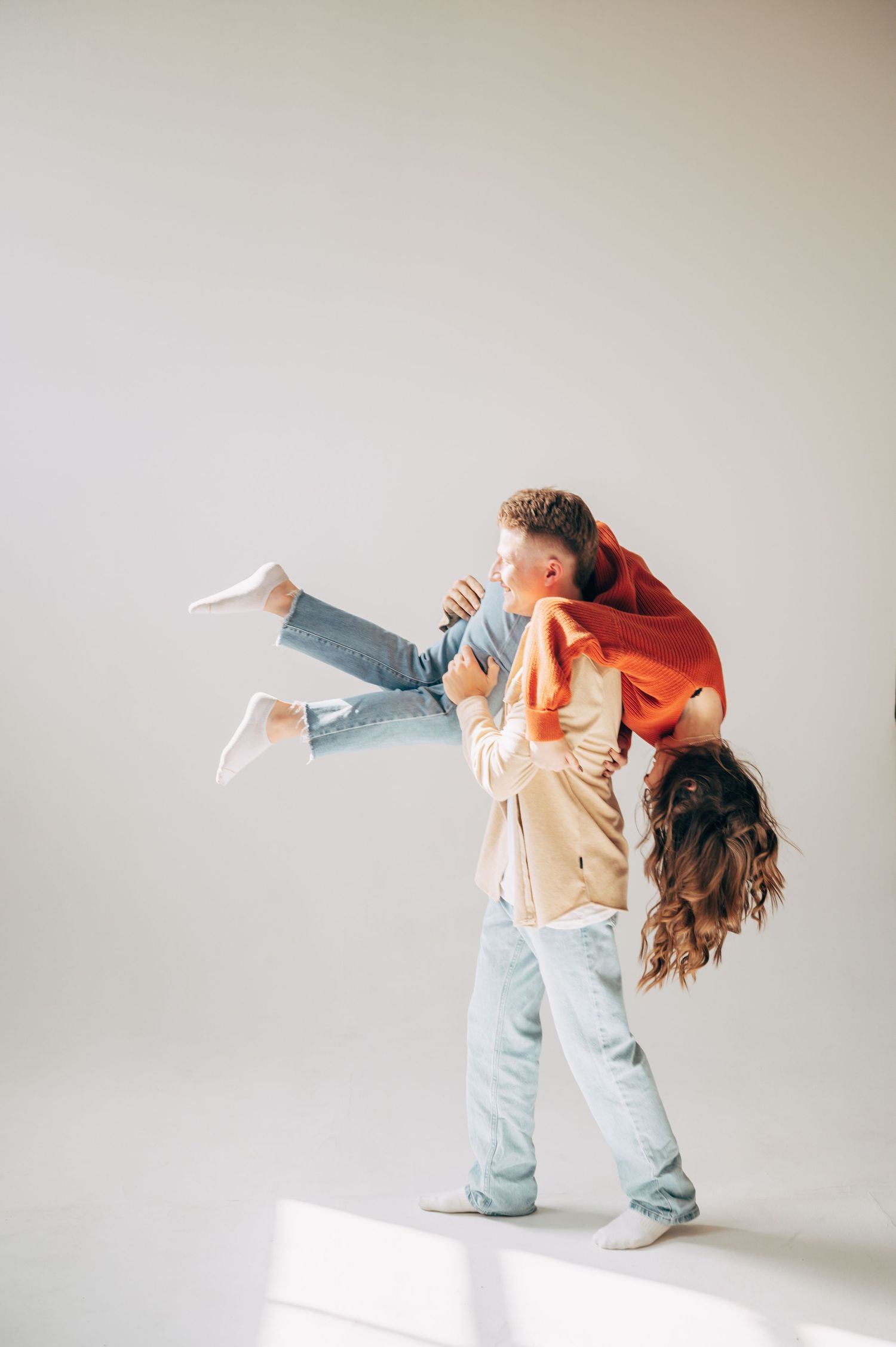 A man in jeans lifts a woman in a red top and jeans, her hair flowing down, against a white background.
