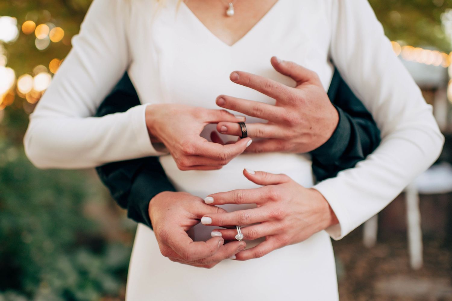Newlywed couple holding hands showing wedding rings, bride in white dress embracing groom outdoors.