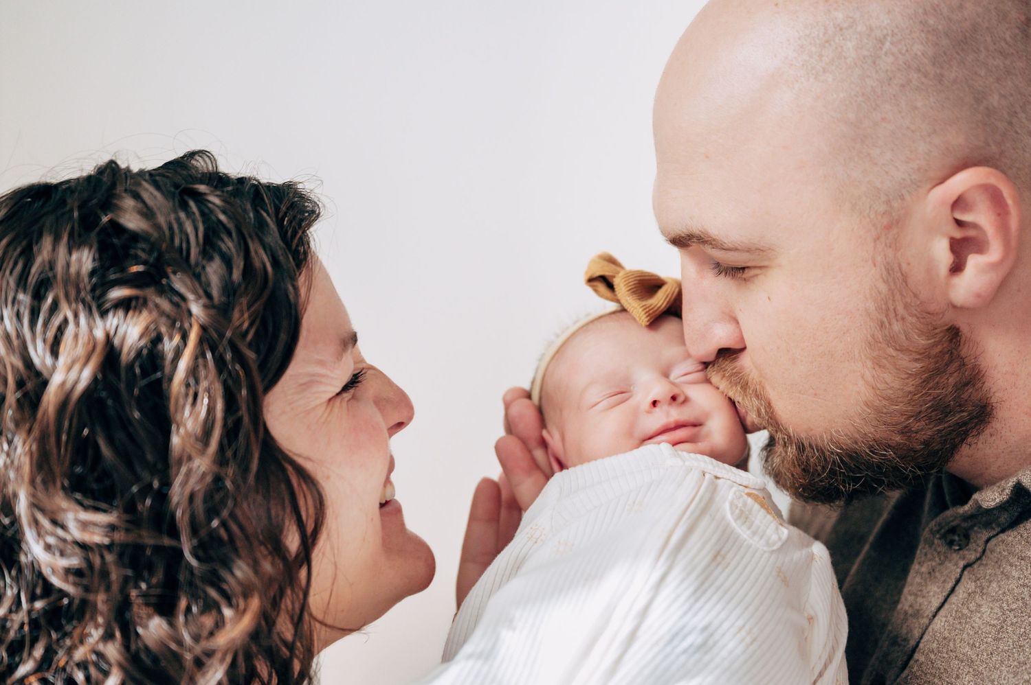 New parents lovingly kiss their newborn baby girl wearing a yellow bow headband against a white background.