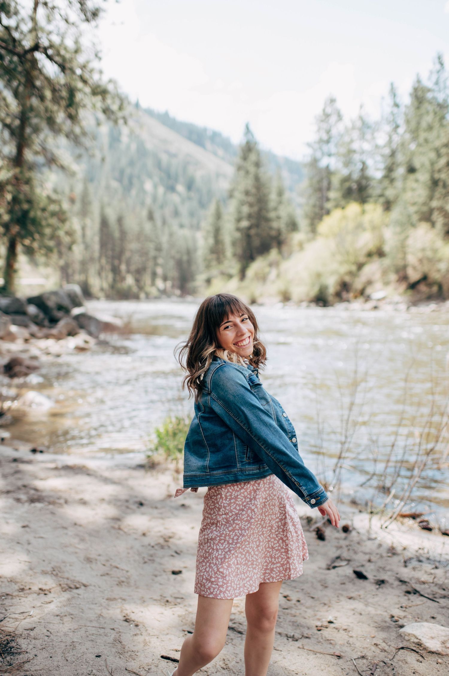 Young woman in denim jacket and floral skirt smiling by a mountain river surrounded by pine trees.