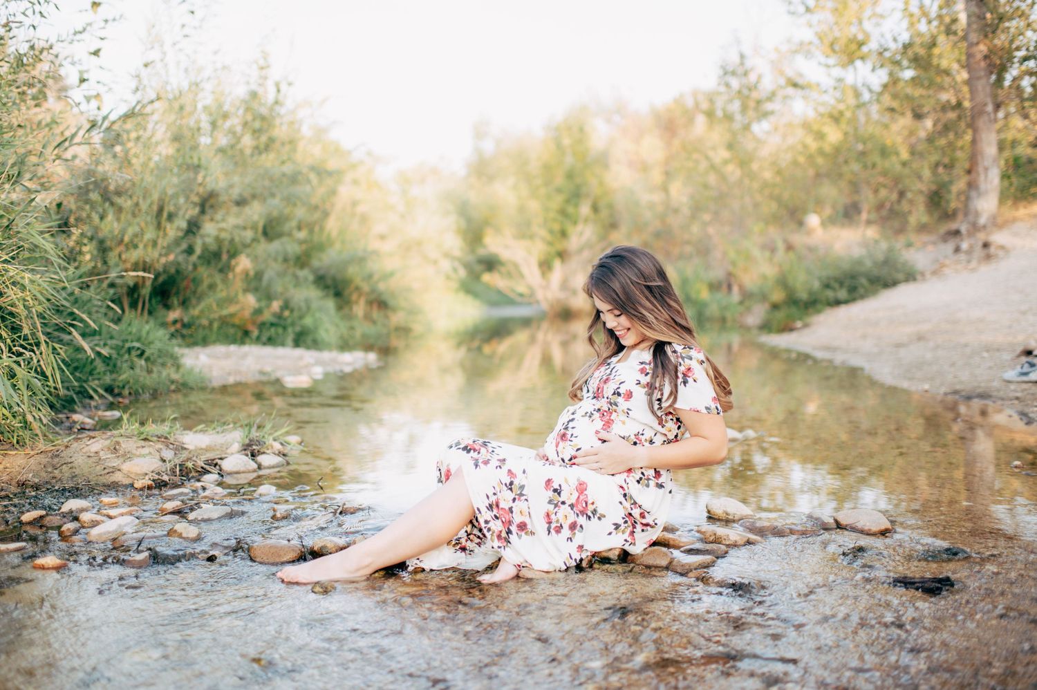 Pregnant woman in floral dress sitting on rocks in a serene creek surrounded by lush green trees.