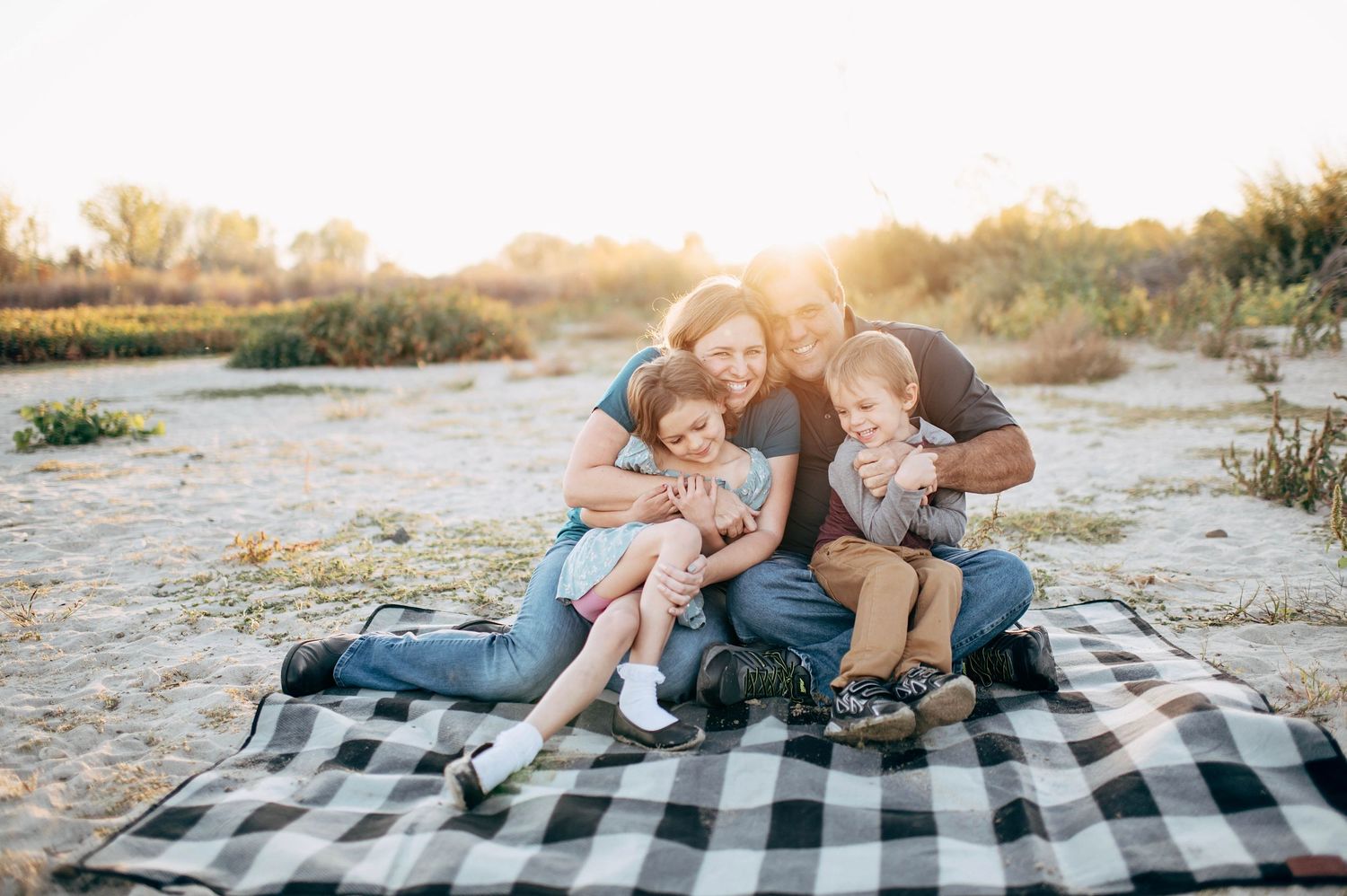 Family of four sitting on plaid blanket outdoors at golden hour, parents hugging two young children in sunny field.