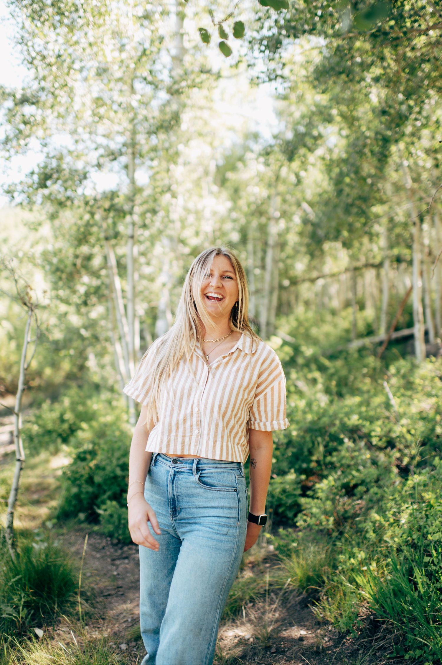 A smiling blonde woman in a striped crop top and jeans stands on a forest path surrounded by tall trees.