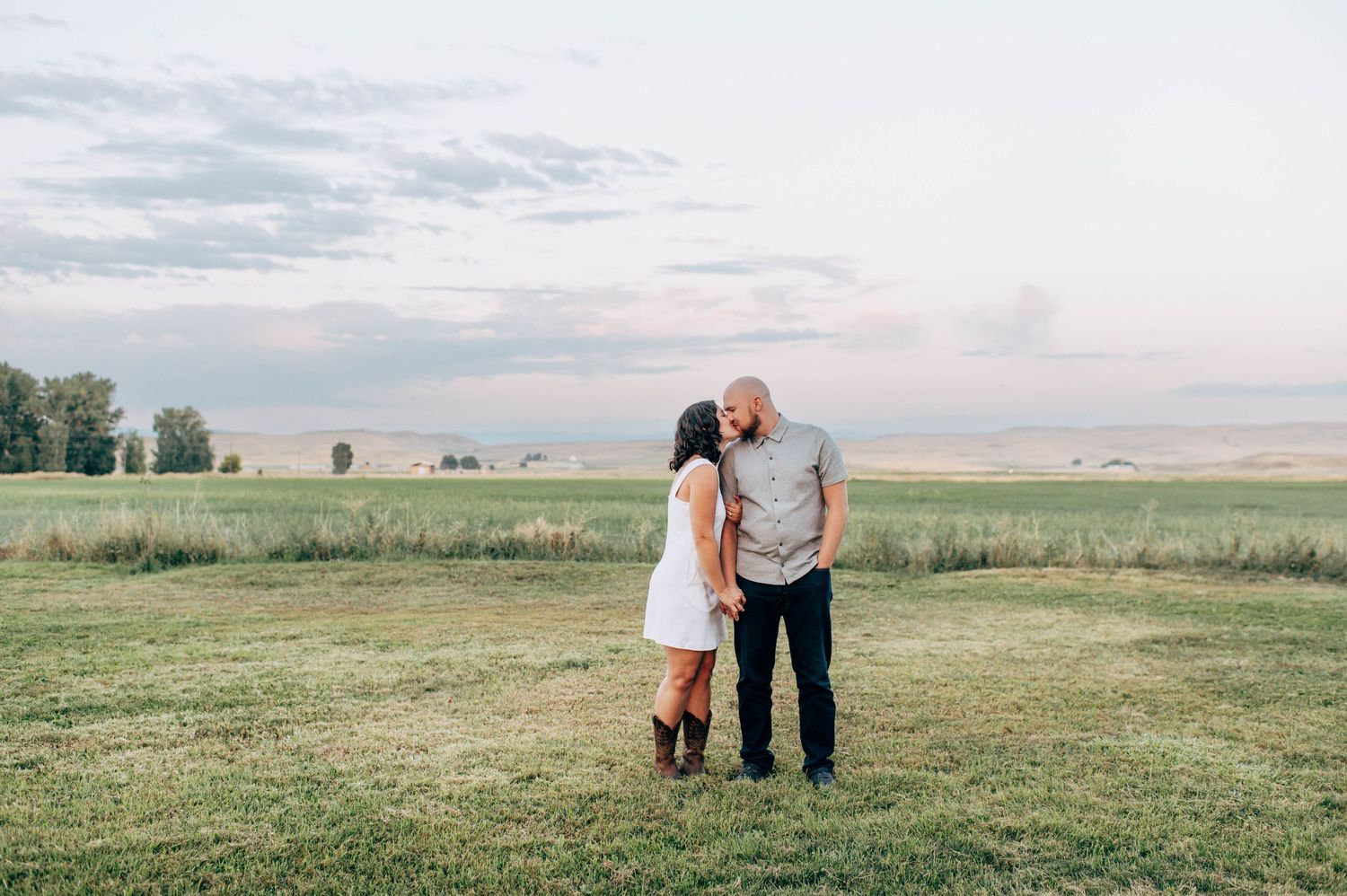 Couple sharing a romantic moment in a green field at sunset with a scenic rural landscape in the background.