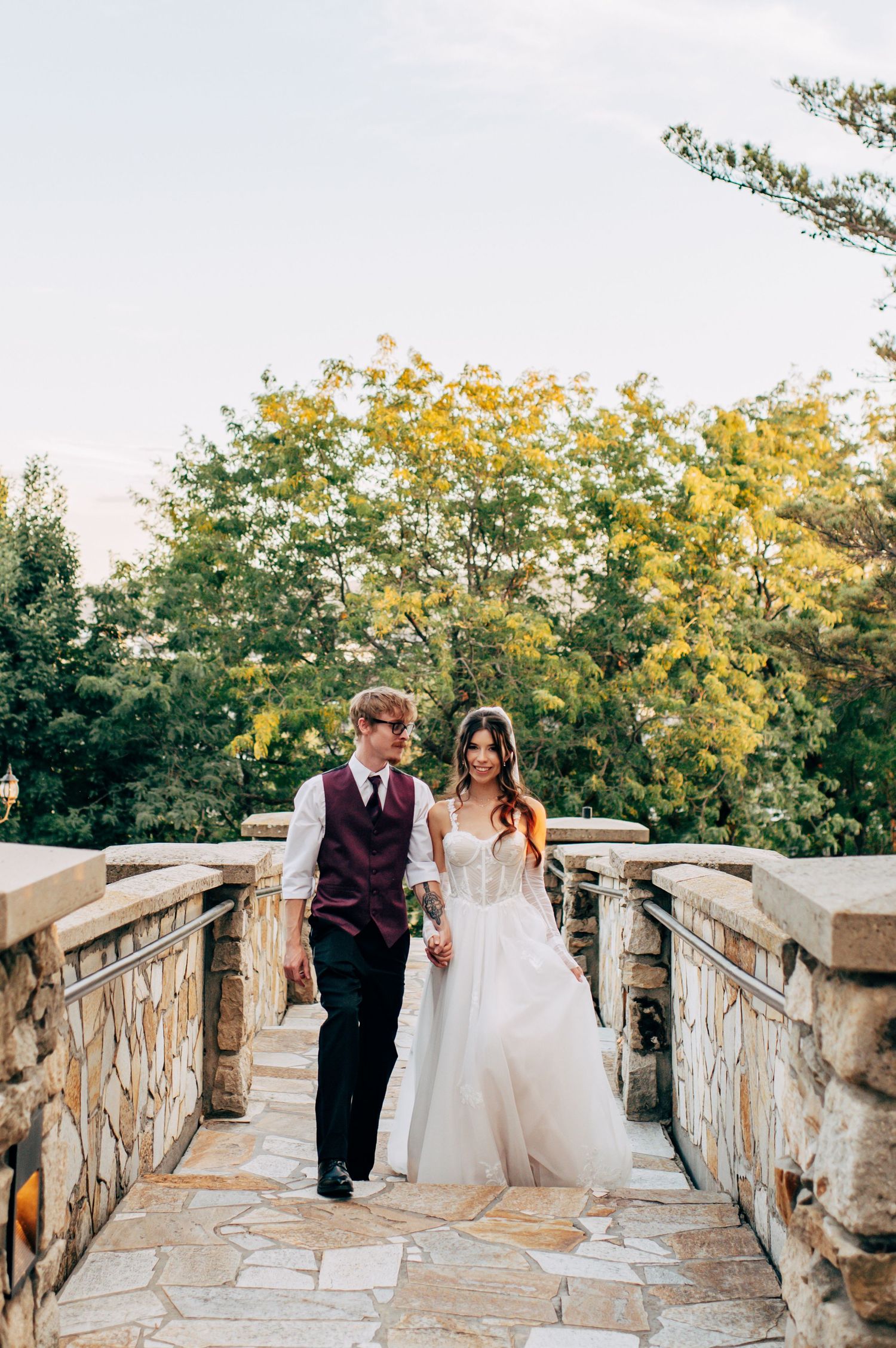 Bride in white gown and groom in burgundy vest walk hand-in-hand on stone bridge surrounded by lush green trees.