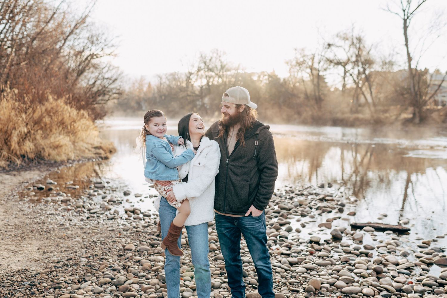 Family of three posing by a rocky riverbank surrounded by bare trees in a warm autumn outdoor portrait.