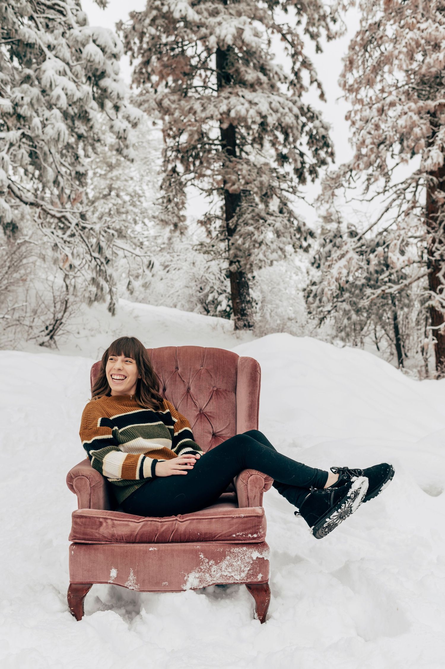 Woman laughing in pink velvet armchair surrounded by snow-covered pine trees, wearing striped sweater and winter boots.