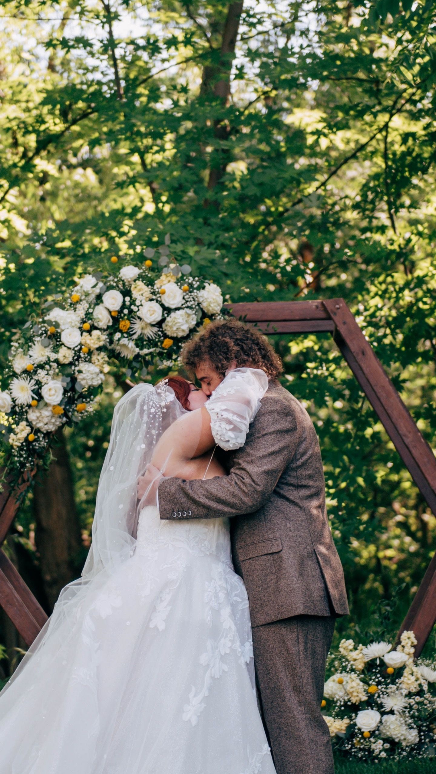 Couple sharing first kiss under wooden arch decorated with white flowers and greenery.