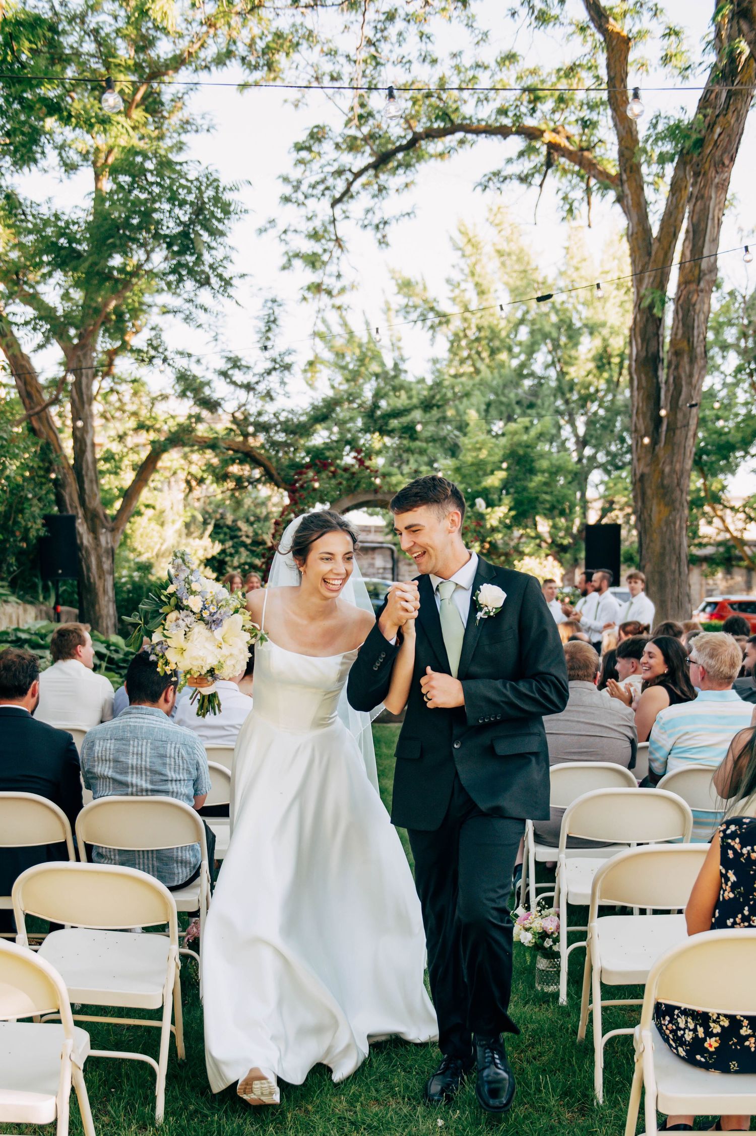 A joyful wedding moment captured outdoors with guests seated in white chairs under mature trees.