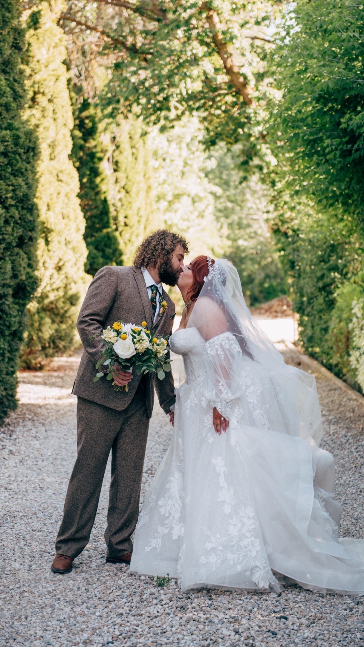 Newlywed couple sharing a romantic moment on a tree-lined gravel path while holding floral bouquet.