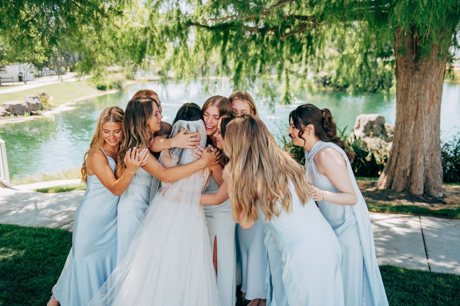 Bride in white gown hugged by bridesmaids in light blue dresses outdoors near a pond under trees.
