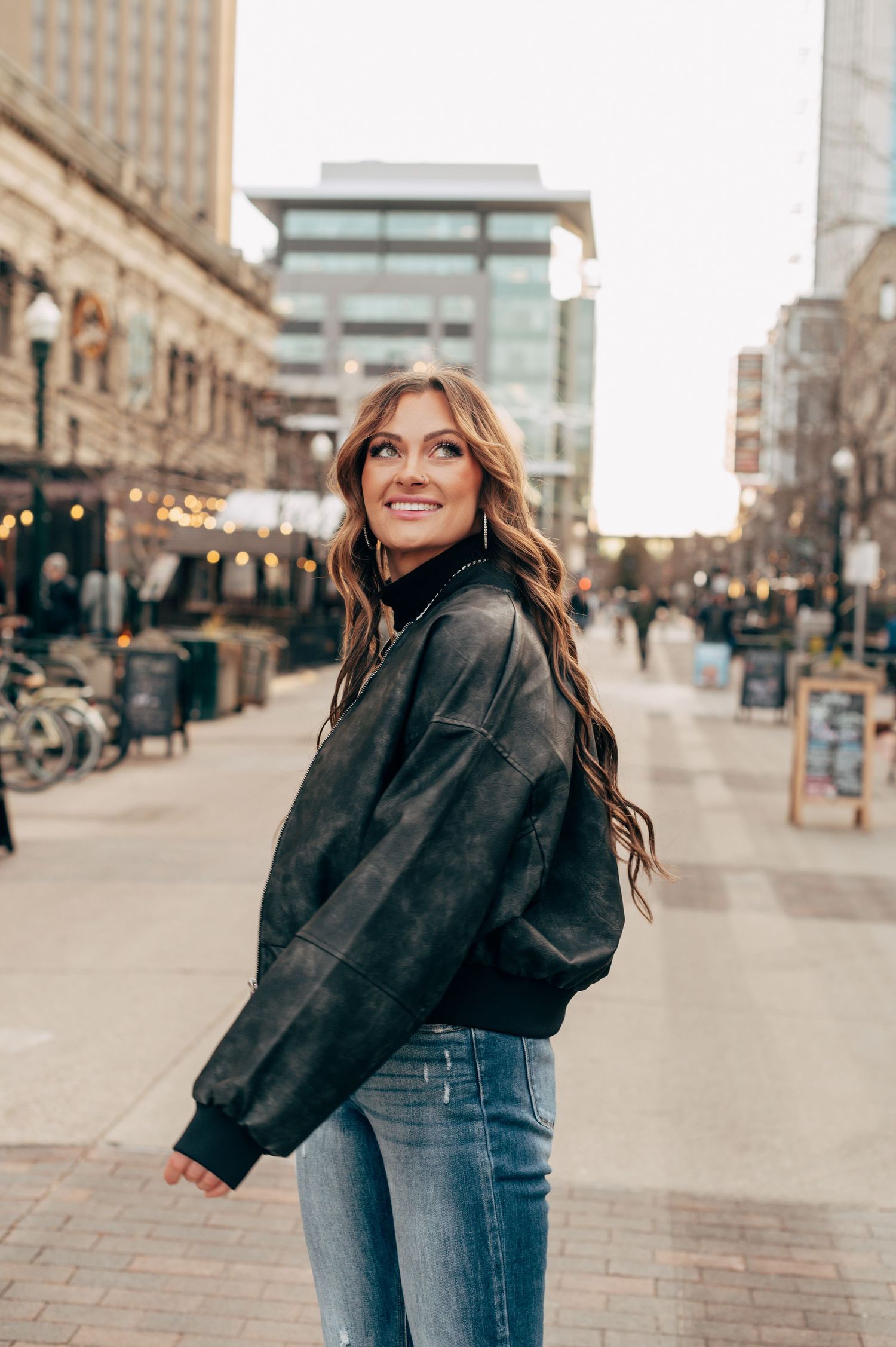 Young woman in black leather bomber jacket and jeans smiling on urban pedestrian street with city buildings at dusk.